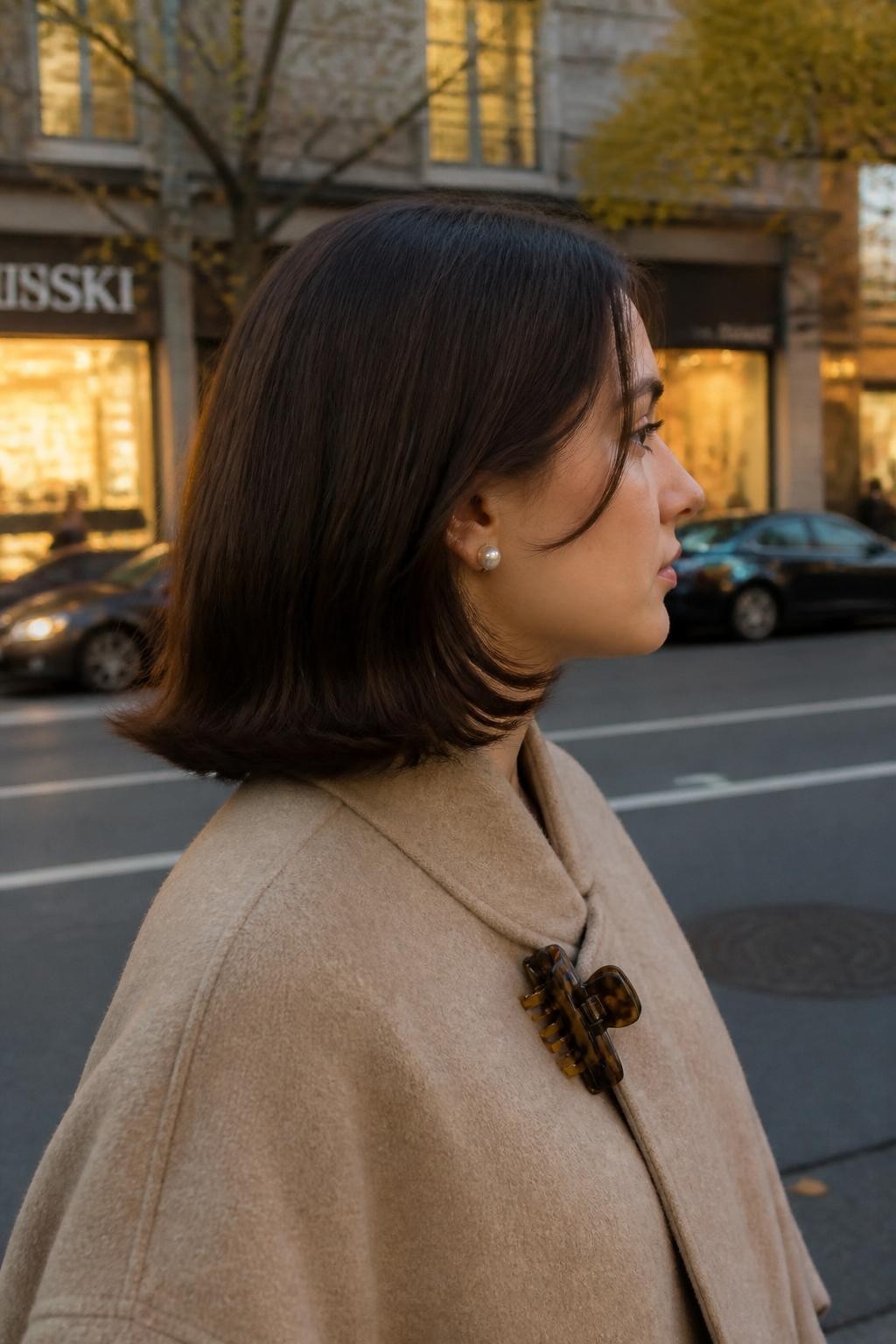 Image prompt: Photo of a woman wearing a shoulder-length bob with smooth roots and evenly flipped-out ends, side profile view, softly blurred city street background with warm shop windows, golden hour glow, containing pearl earrings peeking through and a claw clip attached to her coat collar, realistic candid fall moment, latest iPhone photo quality.