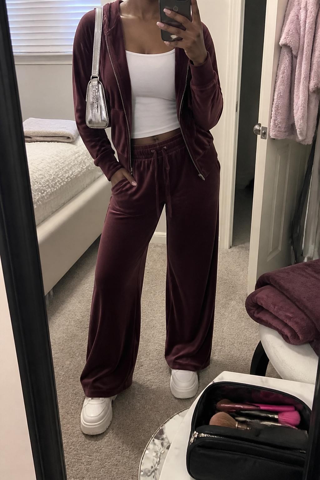 mirror selfie of a Black woman wearing a burgundy velour zip hoodie, matching burgundy velour wide-leg pants, white fitted tank top, white platform sneakers, and a silver mini shoulder bag, clearly showing the same clothing items, colors, fabrics, and accessories listed in the Outfit Pieces section, holding a phone in front of a mirror, cropped at NECK so the face does not show, with a relaxed casual selfie pose, in a cozy bedroom mirror setup, soft indoor lighting, including a folded blanket on a chair and a makeup brush or cosmetic bag on the counter, latest iPhone photo quality.