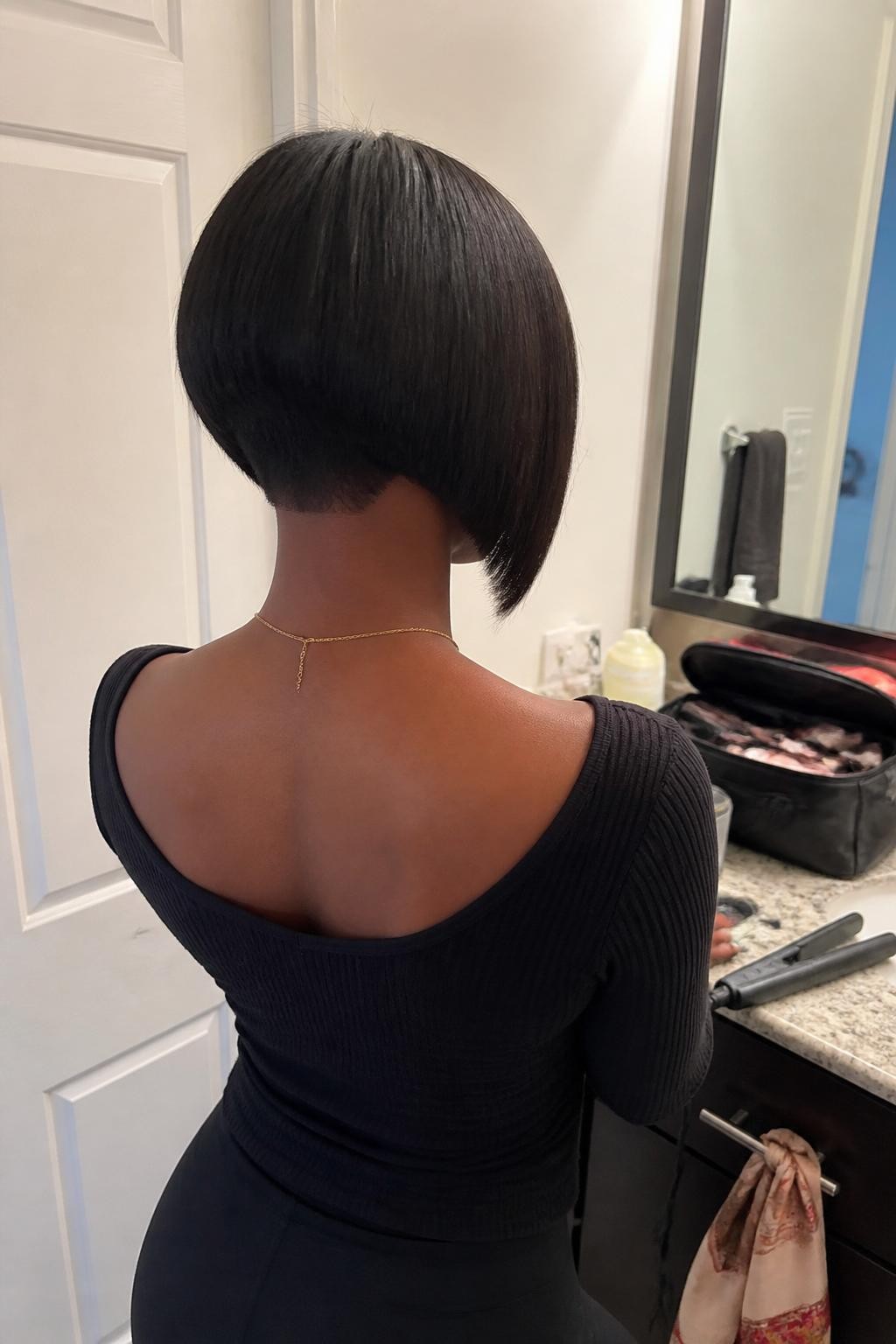 Image prompt: Photo of a Black woman wearing a sleek angled bob that is cropped at the nape and longer near the chin, back-of-head view to highlight the graduated cut, bathroom vanity mirror with a makeup bag nearby, soft indoor lighting, containing a flat iron on the counter and a satin scarf hanging from a drawer handle, realistic getting-ready moment, latest iPhone photo quality.
