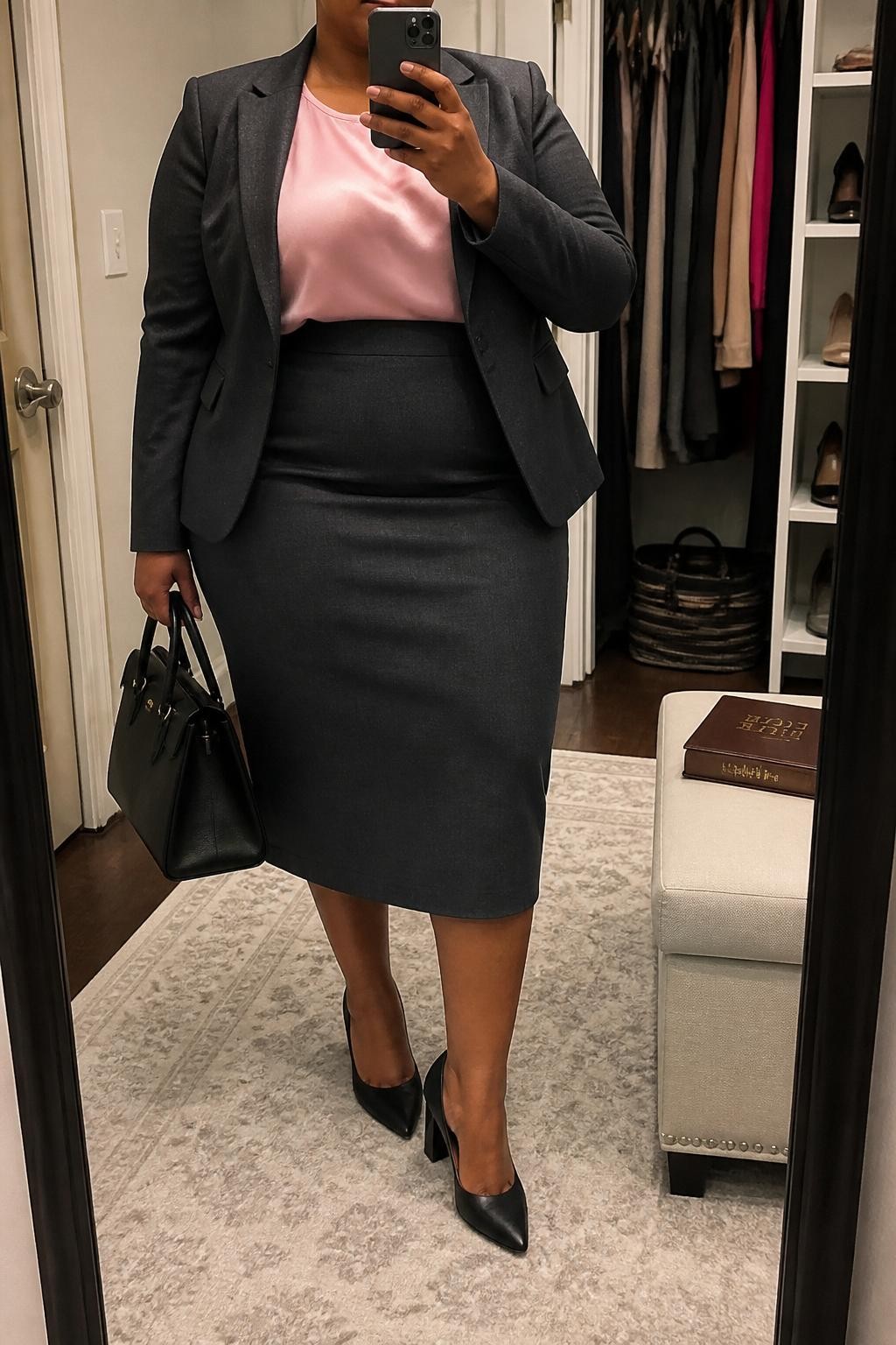 Mirror selfie of a plus size woman wearing a charcoal grey tailored blazer with a matching high-waisted pencil midi skirt and back walking slit, a fitted blush pink satin shell top with a modest round neckline, black pointed block-heel pumps, and a structured black handbag, clearly showing the exact suit, charcoal fabric, blush satin, footwear, and accessories listed, holding a phone in front of a mirror, cropped at the neck so her face is hidden, relaxed professional selfie stance, in a walk-in closet mirror with soft indoor lighting, an open closet with clothes visible and a Bible resting on a bench, realistic formal church outfit, latest iPhone photo quality.