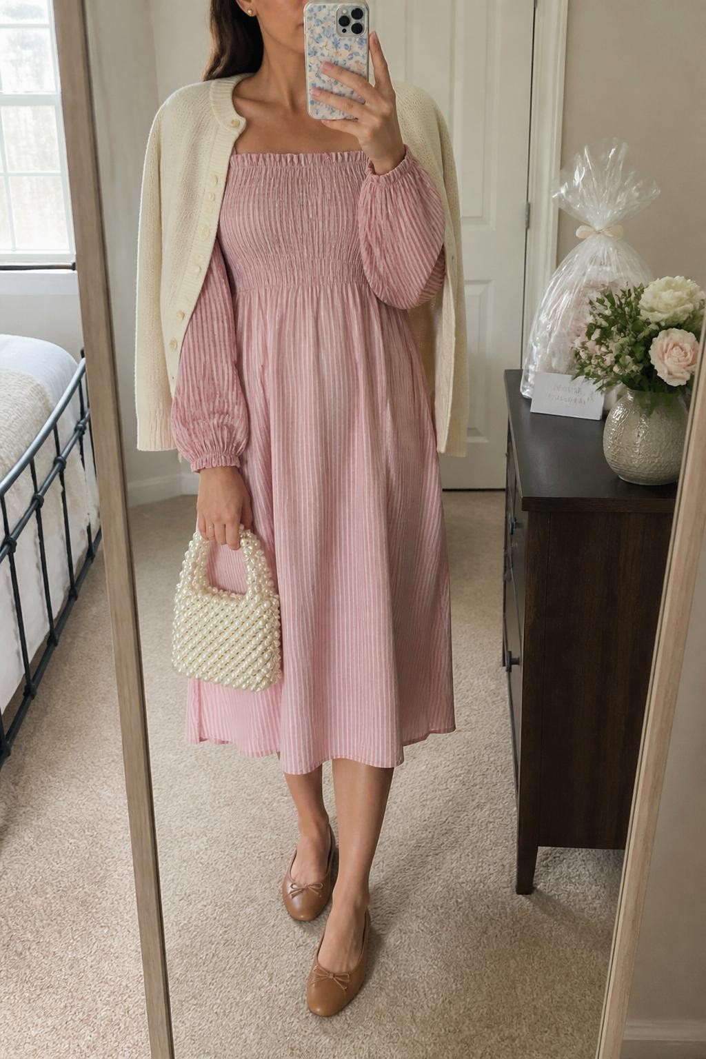 mirror selfie of a woman wearing the exact outfit described above, clearly showing a blush pink smocked midi dress with long puff sleeves, tan leather ballet flats, a cream cardigan draped over the shoulders, a pearl-beaded handbag, and small gold stud earrings, holding a phone in front of a mirror, cropped at NECK so the face does not show, with a relaxed casual selfie pose, in a bright guest room mirror setup, natural daylight, with a small bouquet and wrapped baby shower gift on the dresser, latest iPhone photo quality.