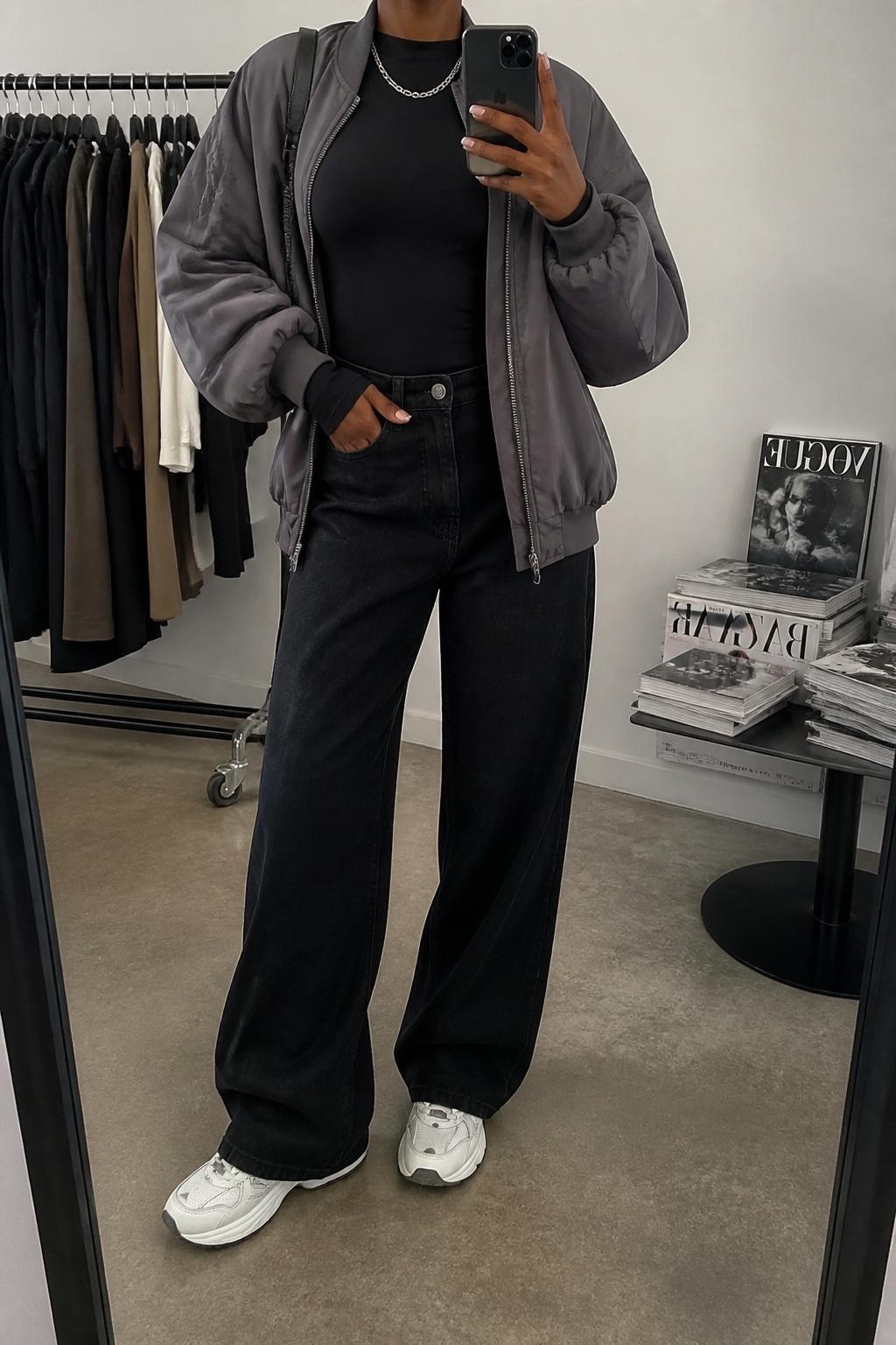 Mirror selfie of a Black woman wearing a gray oversized bomber jacket, fitted black long-sleeve top, black wide-leg jeans, white chunky sneakers, silver chain necklace, and a black mini shoulder bag, holding a phone in front of a mirror, cropped at NECK so the face does not show, relaxed casual selfie pose, in a fashion studio mirror corner, soft indoor lighting, with a clothing rack with garments behind and a stack of fashion magazines on a side table, latest iPhone photo quality.