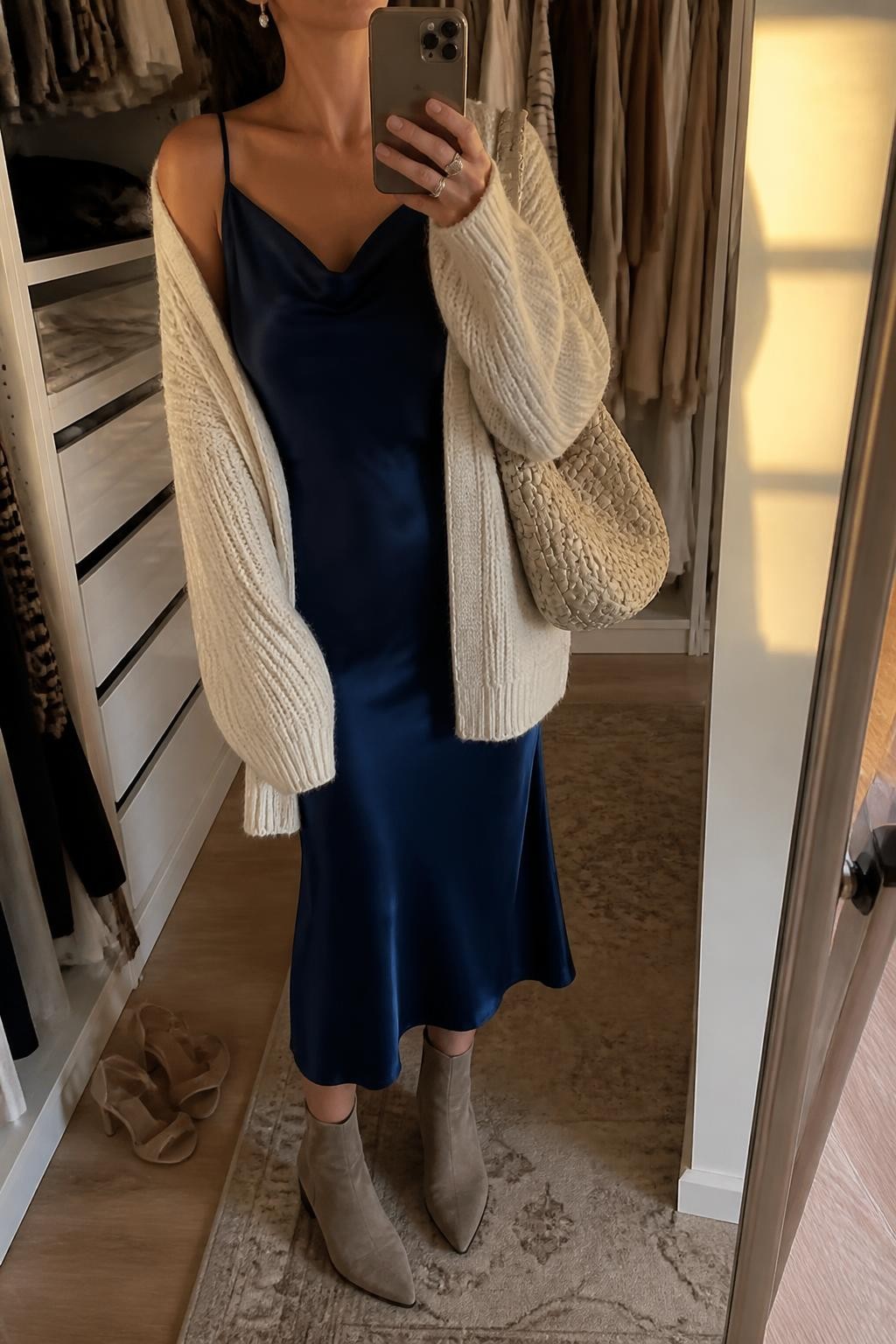 Mirror selfie of a woman wearing a navy satin midi slip dress, oversized cream chunky-knit cardigan, taupe suede ankle boots, a woven cream shoulder bag, and small pearl drop earrings, clearly showing the exact clothing items, colors, fabrics, and accessories listed, holding a phone in front of a walk-in closet mirror, cropped at the neck so the face does not show, relaxed casual selfie pose, evening golden-hour lighting, an open closet with clothes visible and a pair of heels on the floor nearby, warm realistic setting, latest iPhone photo quality.
