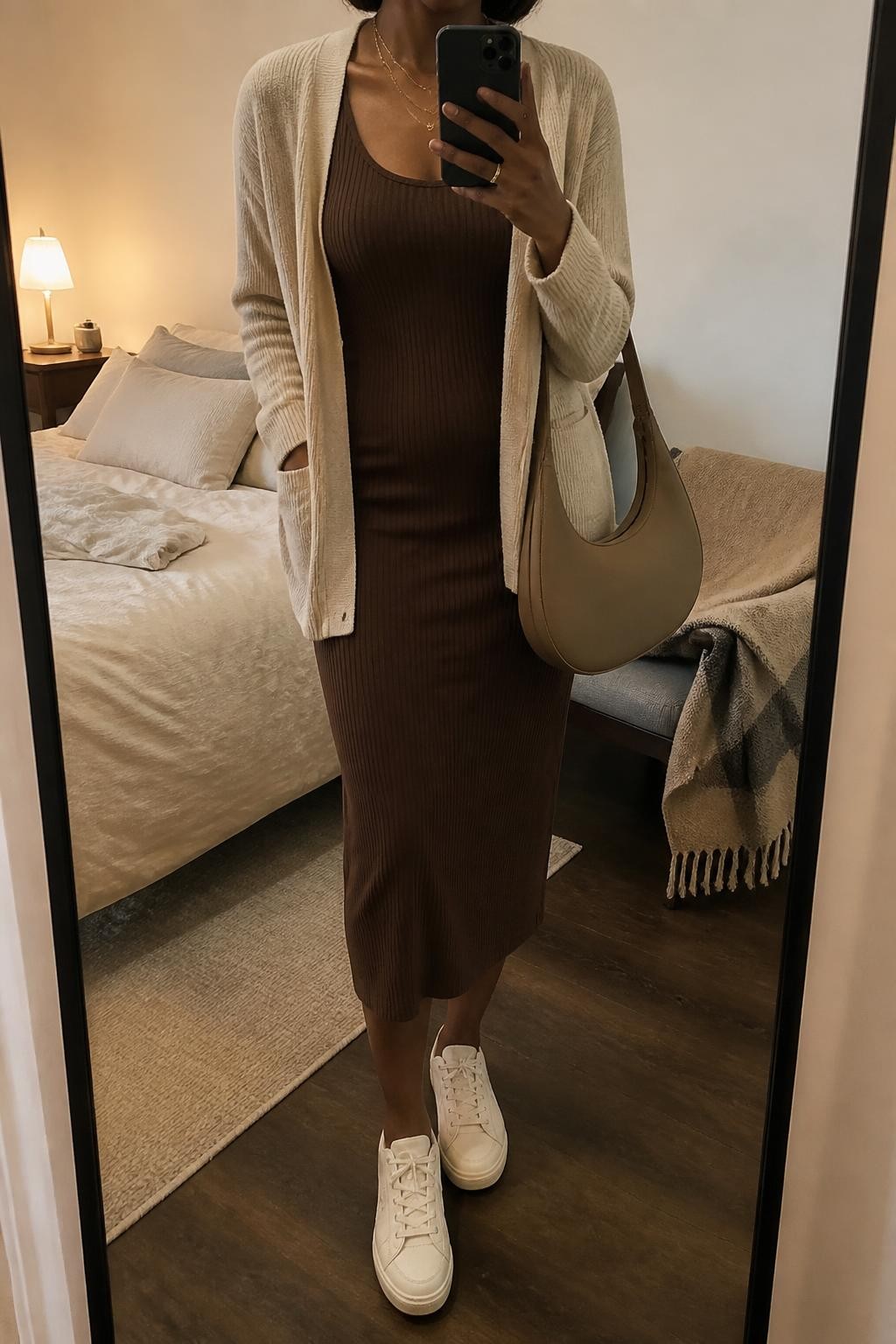 Mirror selfie of a Black woman wearing a fitted chocolate-brown ribbed tank midi dress, lightweight oatmeal open-front cardigan, cream leather sneakers, and a soft tan hobo bag, clearly showing the exact clothing items, colors, fabrics, and accessories listed, holding a phone in front of a cozy bedroom mirror setup, cropped at the neck so the face does not show, relaxed casual selfie pose, warm moody lighting, soft pillows on an unmade bed and a folded blanket on a chair, realistic and uncluttered, latest iPhone photo quality.