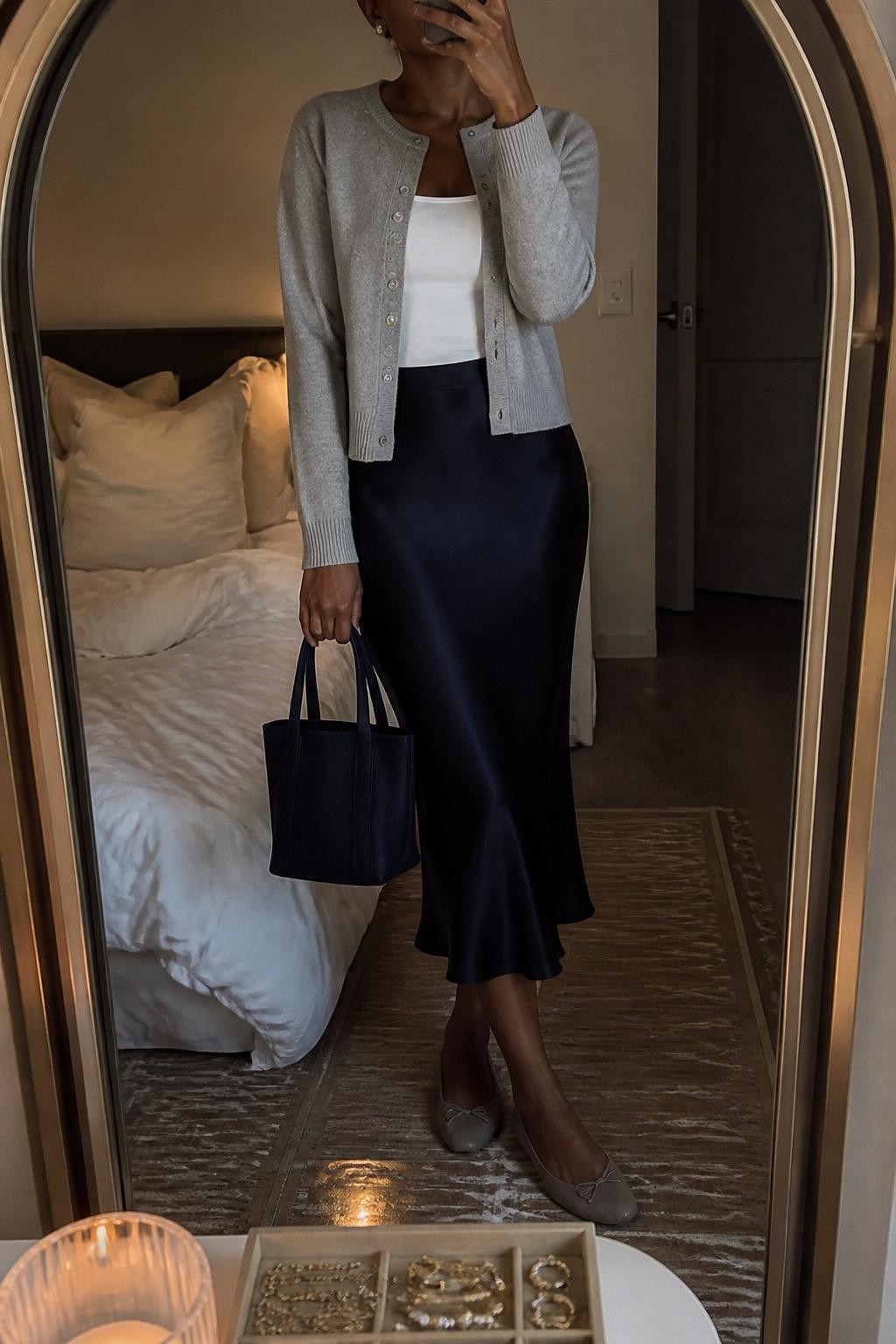 Mirror selfie of a Black woman wearing a soft gray button-front cardigan, navy satin midi skirt, white fitted tank top, gray suede ballet flats, pearl studs, and a navy mini tote bag, cropped at neck so the face does not show, in a cozy bedroom mirror setup with warm moody lighting, soft pillows on an unmade bed and a jewelry tray on the vanity, realistic latest iPhone photo quality.