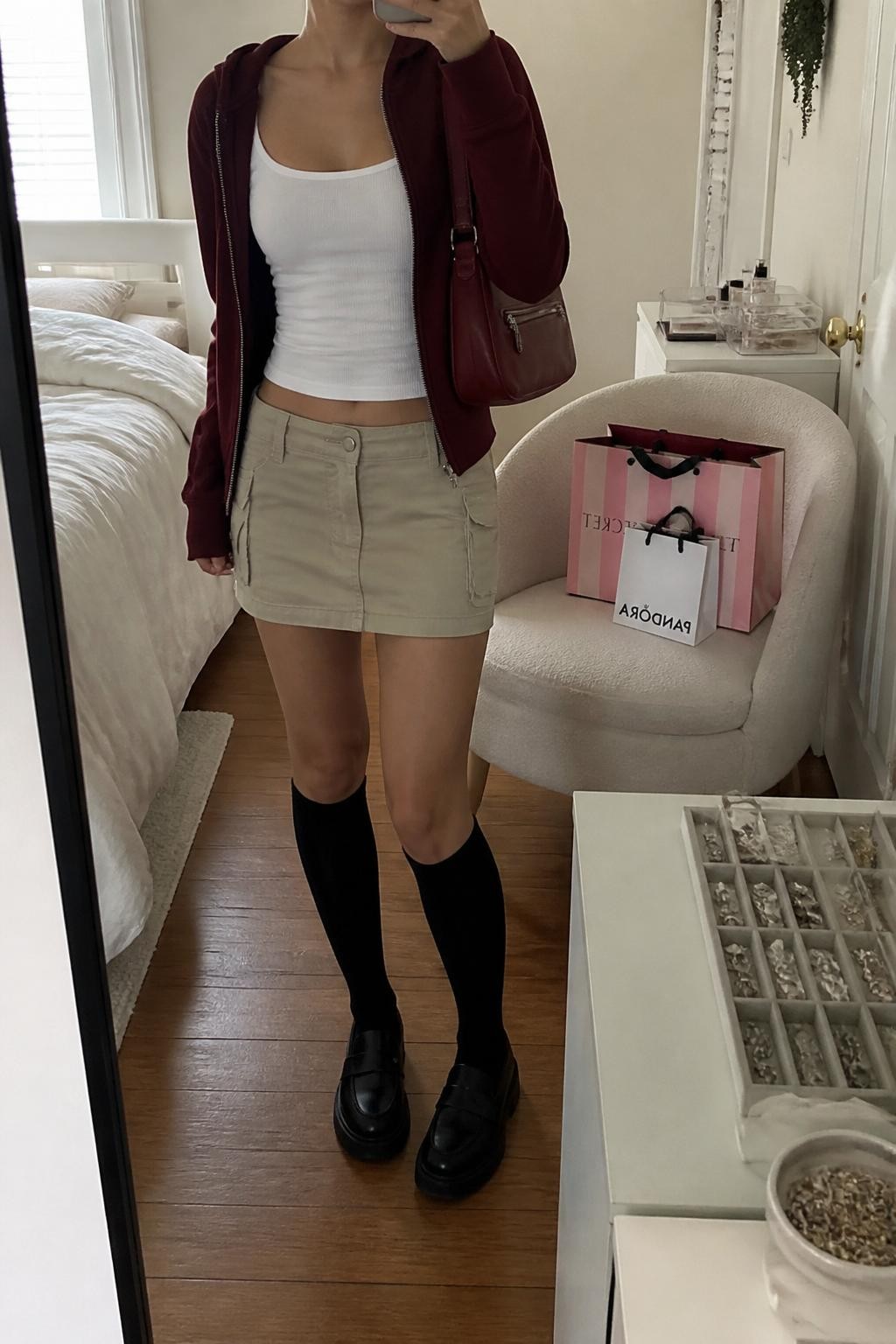 Mirror selfie of a woman wearing a burgundy zip hoodie, white fitted tank top, beige cargo mini skirt, black knee-high socks, black chunky loafers, and a burgundy nylon shoulder bag, holding a phone in front of a mirror, cropped at NECK so the face does not show, relaxed casual selfie pose, in a cozy bedroom mirror setup, natural window light, with shopping bags beside a chair and a jewelry tray on the vanity, latest iPhone photo quality.