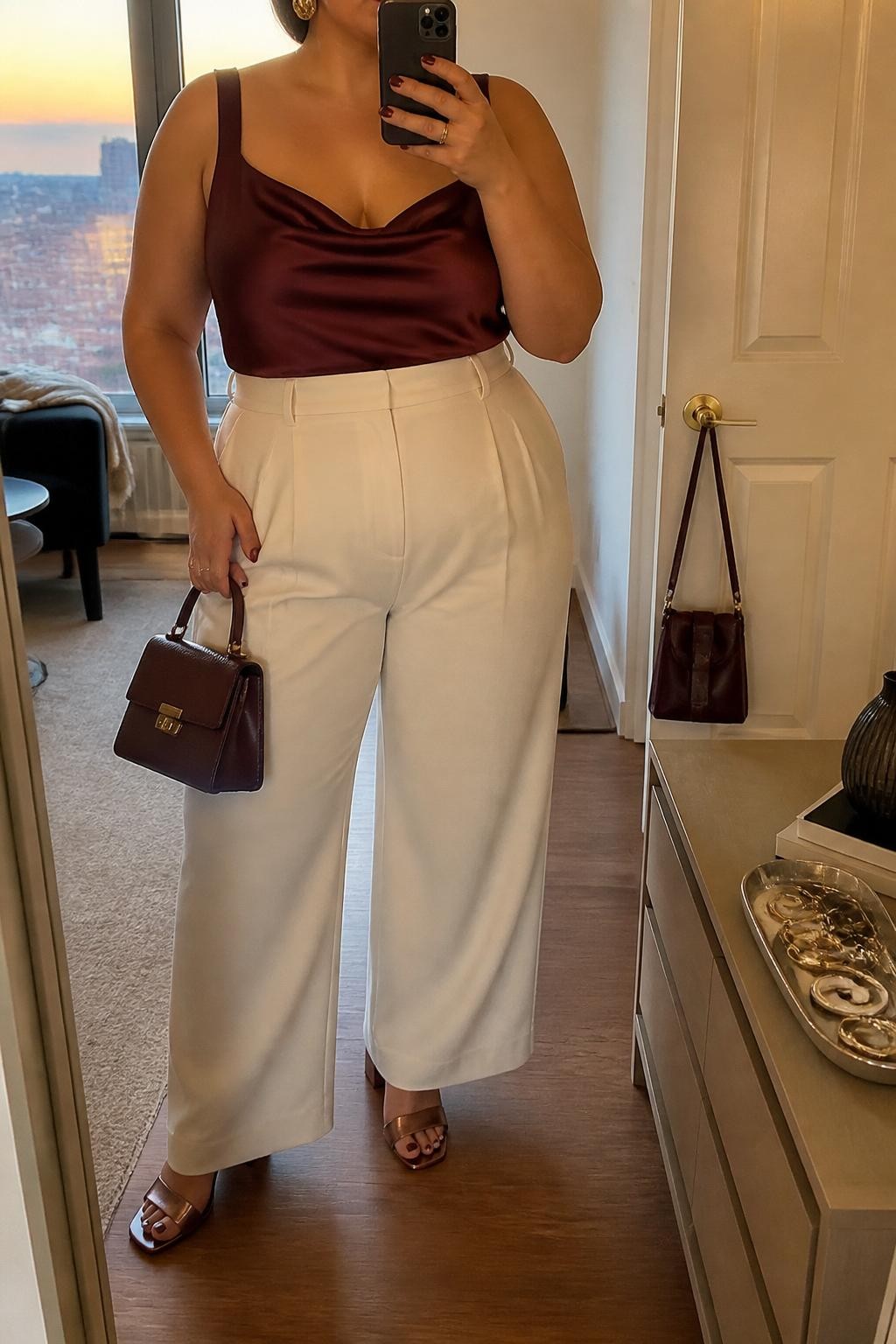 Mirror selfie of a plus size woman wearing cream high-waisted wide-leg trousers, a burgundy satin camisole with a softly draped neckline and wide supportive straps, metallic bronze block-heel sandals, a structured burgundy mini handbag, and hammered gold earrings, clearly showing the exact clothing items, cream tailoring, burgundy satin, footwear, and accessories listed, holding a phone in front of a mirror, cropped at the neck so the face does not appear, relaxed elegant selfie pose, in a stylish city apartment with evening golden-hour lighting, a jewelry tray on the dresser and a handbag hanging on a door handle, realistic rooftop-date outfit, latest iPhone photo quality.