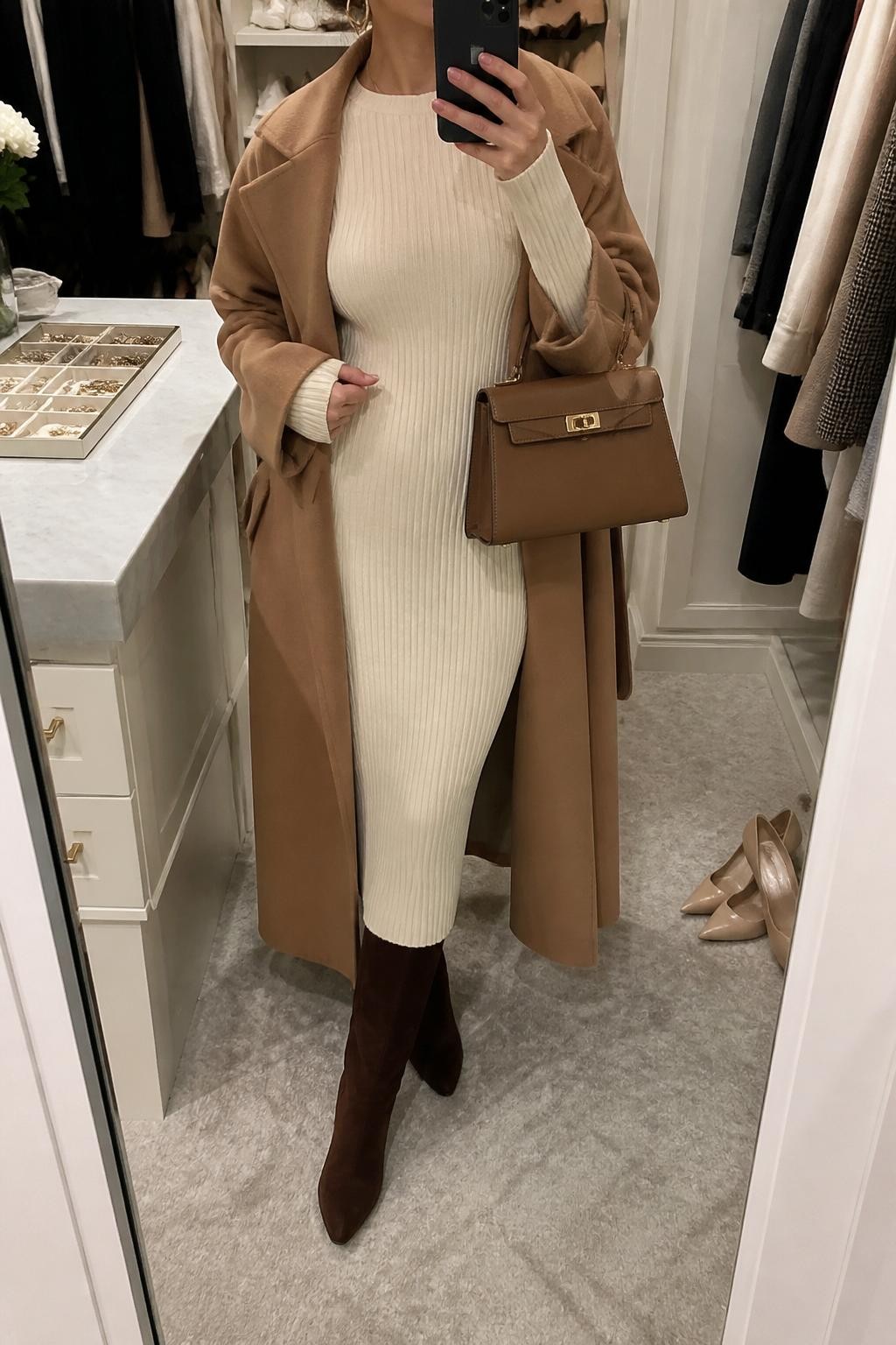 mirror selfie of a woman wearing the exact outfit described above, clearly showing chocolate brown suede knee-high boots, a cream ribbed sweater midi dress with long sleeves, a camel wrap coat worn open, a tan structured handbag, and gold hoop earrings, holding a phone in front of a mirror, cropped at NECK so the face does not show, with a relaxed casual selfie pose, in a walk-in closet mirror, soft indoor lighting, with a jewelry tray on the vanity and a pair of heels on the floor nearby, latest iPhone photo quality.