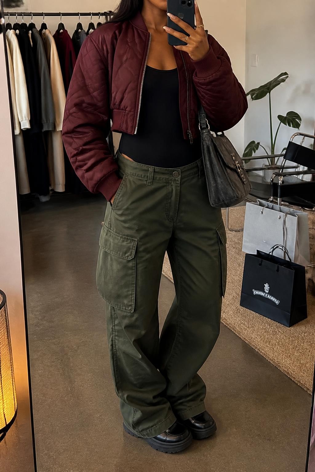 Mirror selfie of a Black woman wearing loose olive-green cargo pants, a fitted black long-sleeve bodysuit, a cropped burgundy quilted bomber jacket, chunky black leather sneakers, and a charcoal-grey messenger bag with silver buckles, clearly showing the exact clothing items, colors, fabrics, and accessories listed, holding a phone in front of a fashion studio mirror corner, cropped at the neck so the face does not show, relaxed casual selfie pose, warm moody lighting, a clothing rack with garments behind and shopping bags beside a chair, casual realistic atmosphere, latest iPhone photo quality.