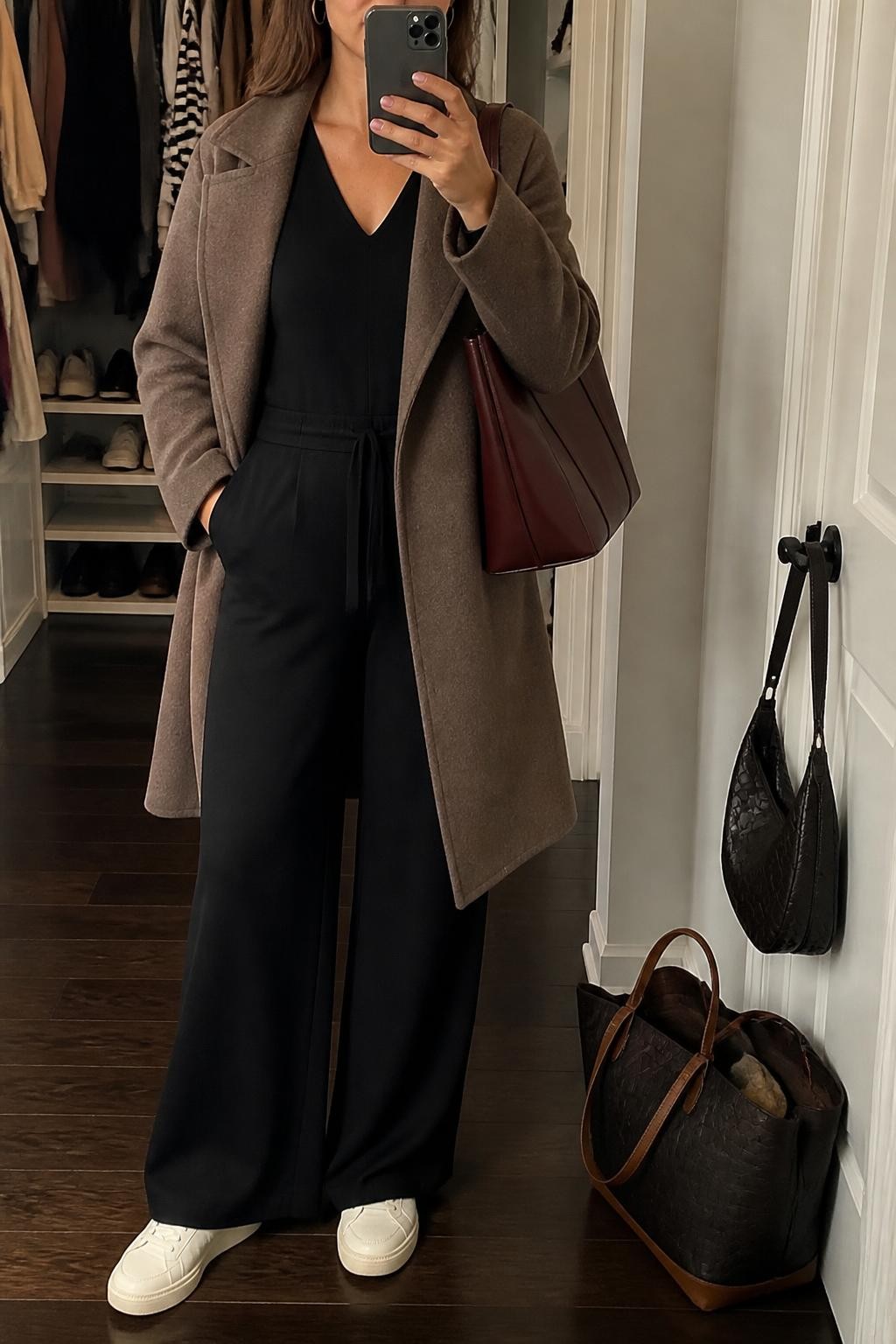 Mirror selfie of a woman wearing a black wide-leg travel jumpsuit in soft stretch fabric, a knee-length mocha wool wrap coat, cream leather sneakers, a burgundy structured tote bag, and small silver hoop earrings, clearly showing the exact clothing items, colors, fabrics, and accessories listed, holding a phone in front of a walk-in closet mirror, cropped at the neck so the face does not show, relaxed casual selfie pose, warm moody lighting, a handbag hanging on a door handle and a tote bag resting on the floor, realistic and uncluttered, latest iPhone photo quality.