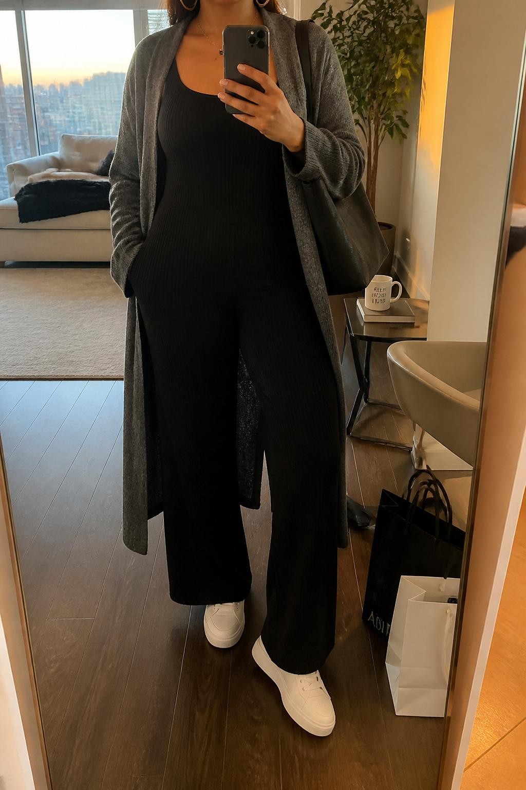 Mirror selfie of a curvy woman wearing a black ribbed wide-leg jumpsuit, long gray coatigan, white low-profile sneakers, silver hoop earrings, and a charcoal tote bag, holding a phone in front of a mirror, cropped at NECK so the face does not show, relaxed casual selfie pose, in a stylish city apartment, evening golden hour, with a coffee mug on a side table and shopping bags beside a chair, latest iPhone photo quality.