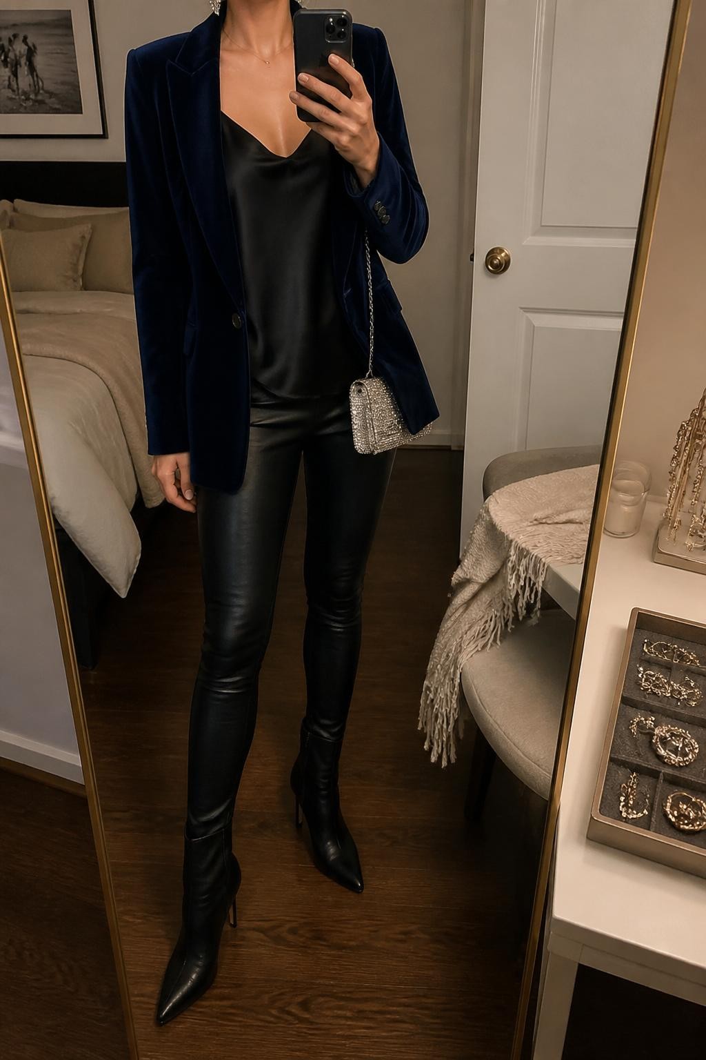 mirror selfie of a woman wearing the exact outfit described above, clearly showing a navy velvet blazer layered over a black satin cami, black leather leggings, black heeled ankle boots, a silver mini bag, and crystal stud earrings, holding a phone in front of a mirror, cropped at NECK so the face does not show, with a relaxed casual selfie pose, in a cozy bedroom mirror setup, warm moody lighting, with a jewelry tray on the vanity and a folded blanket on a chair, latest iPhone photo quality.