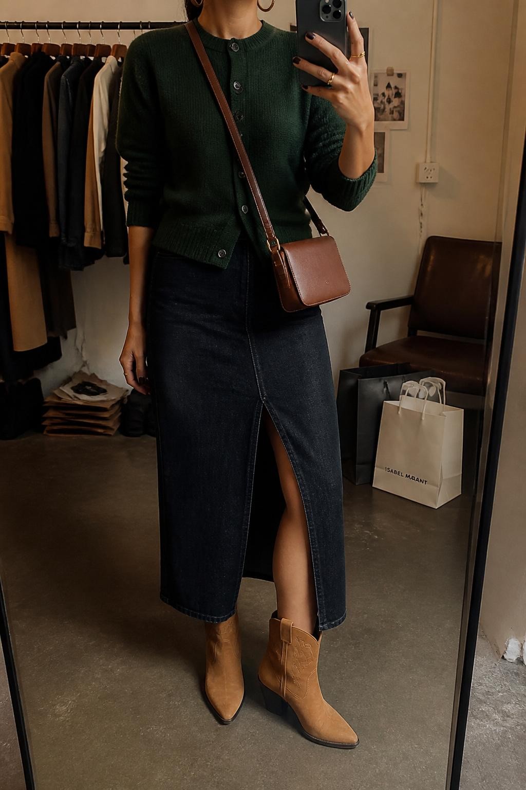 mirror selfie of a woman wearing the exact outfit described above, clearly showing a forest green button-front cardigan, dark denim midi skirt with a front slit, tan western ankle boots, a cognac crossbody bag, and small gold hoop earrings, holding a phone in front of a mirror, cropped at NECK so the face does not show, with a relaxed casual selfie pose, in a fashion studio mirror corner, warm moody lighting, with a clothing rack with garments behind and shopping bags beside a chair, latest iPhone photo quality.