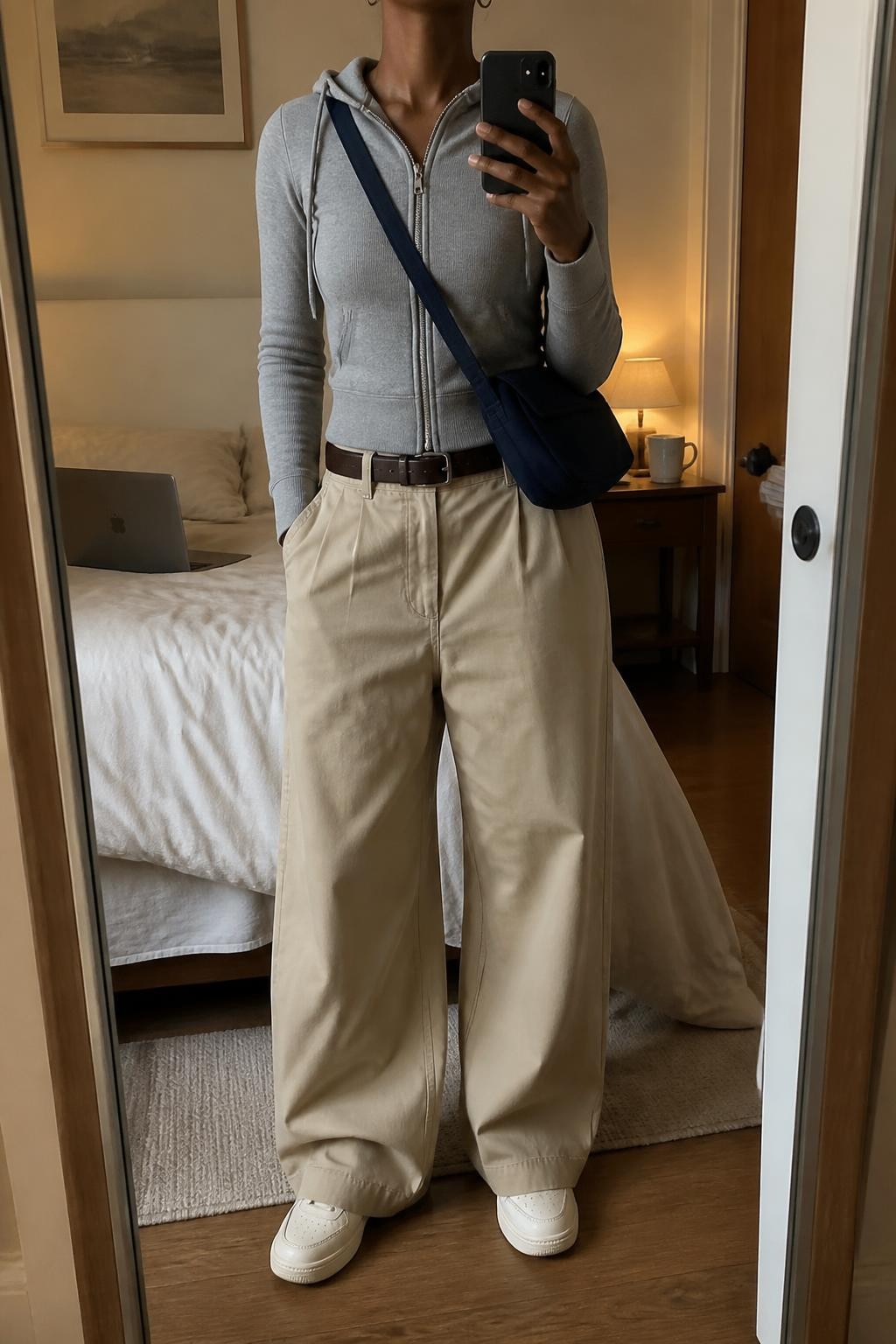 Mirror selfie of a Black woman wearing a soft grey fitted hoodie, sand-beige wide-leg cotton trousers, cream leather sneakers, a navy nylon crossbody bag, and small silver hoop earrings, clearly showing the exact clothing items, colors, fabrics, and accessories listed, holding a phone in front of a cozy bedroom mirror setup, cropped at the neck so the face does not show, relaxed casual selfie pose, warm moody lighting, a laptop on the bed and a coffee mug on a bedside table, casual and realistic, latest iPhone photo quality.