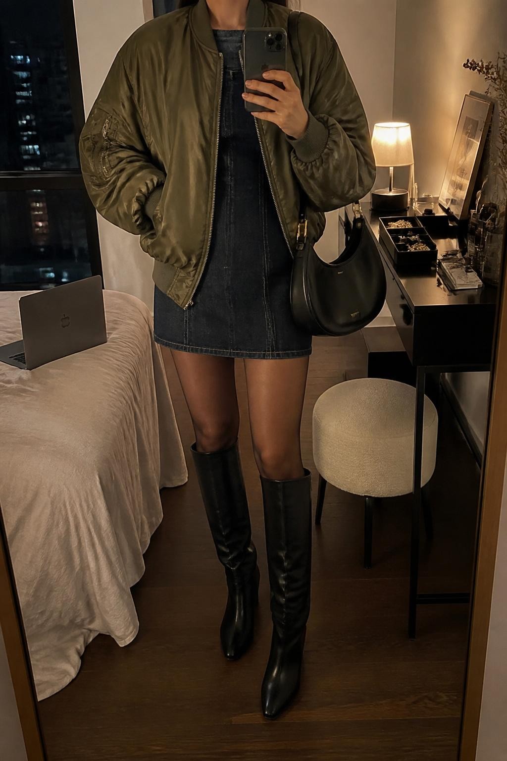 mirror selfie of a woman wearing an olive satin bomber jacket over a dark denim mini dress, sheer black tights, black knee-high boots, and a black crescent shoulder bag, clearly showing the same clothing items, colors, fabrics, and accessories listed in the Outfit Pieces section, holding a phone in front of a mirror, cropped at NECK so the face does not show, with a relaxed casual selfie pose, in a stylish city apartment, warm moody lighting, including a laptop or tablet on the bed and a jewelry tray on the vanity, latest iPhone photo quality.