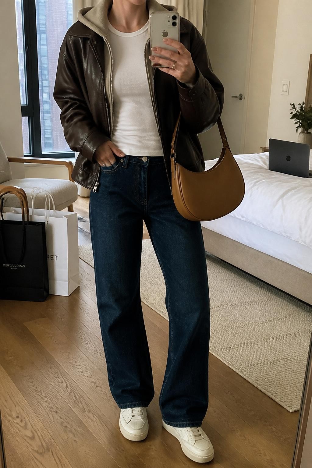 Mirror selfie of a woman wearing a chocolate brown leather jacket over a slim beige hoodie and a fitted white crew-neck T-shirt, clearly showing dark blue straight-leg jeans, cream leather sneakers, and a tan crescent shoulder bag, holding an iPhone in front of a stylish city apartment mirror, cropped at the neck so the face does not show, relaxed casual selfie pose, soft indoor lighting, a laptop on the bed and shopping bags beside a chair, natural unstaged setting, latest iPhone photo quality.