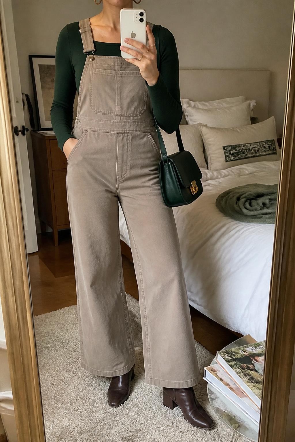 Mirror selfie of a woman wearing taupe wide-leg twill overalls over a fitted forest green square-neck knit top, clearly showing dark brown block-heel boots, a deep green leather handbag, and small brass hoop earrings, holding an iPhone in front of a cozy bedroom mirror setup, cropped at the neck so the face does not show, relaxed casual selfie pose, soft indoor lighting, soft pillows on an unmade bed and a stack of fashion magazines on a side table, natural unstaged setting, latest iPhone photo quality.