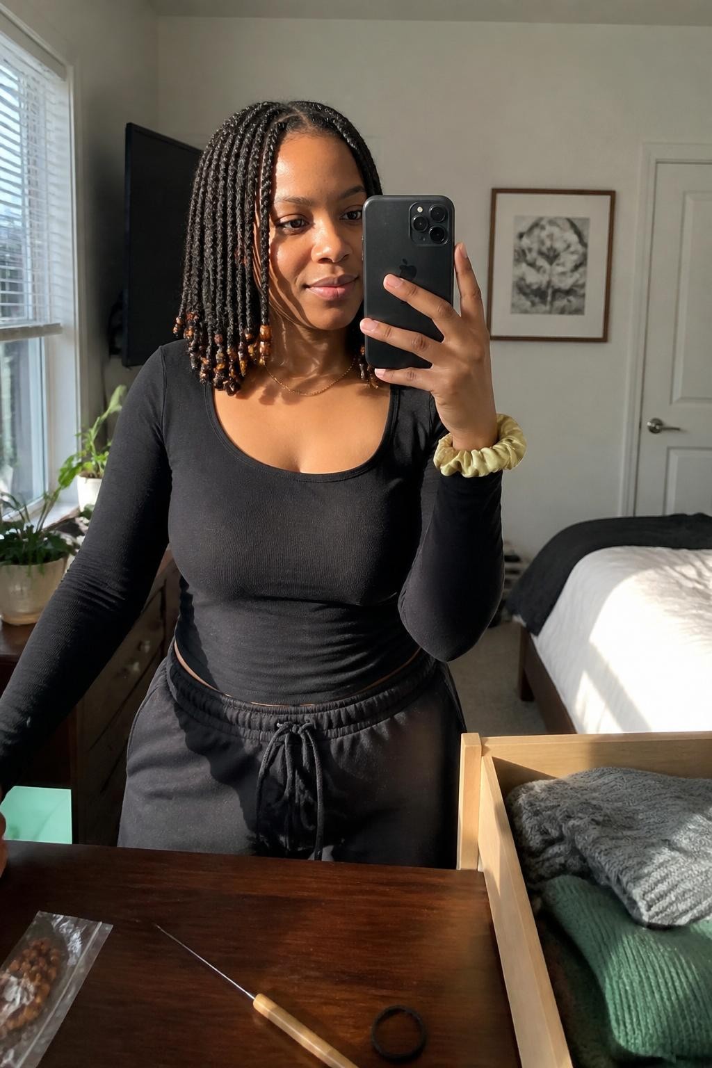 Image prompt: Photo of a Black woman wearing shoulder-length mini twists decorated with a few amber beads at the ends and a neat center part, candid mirror selfie, sun-drenched bedroom with an open drawer and folded sweaters visible, natural window light, containing a beading tool on the dresser and a silk scrunchie around her wrist, realistic home styling moment, latest iPhone photo quality.