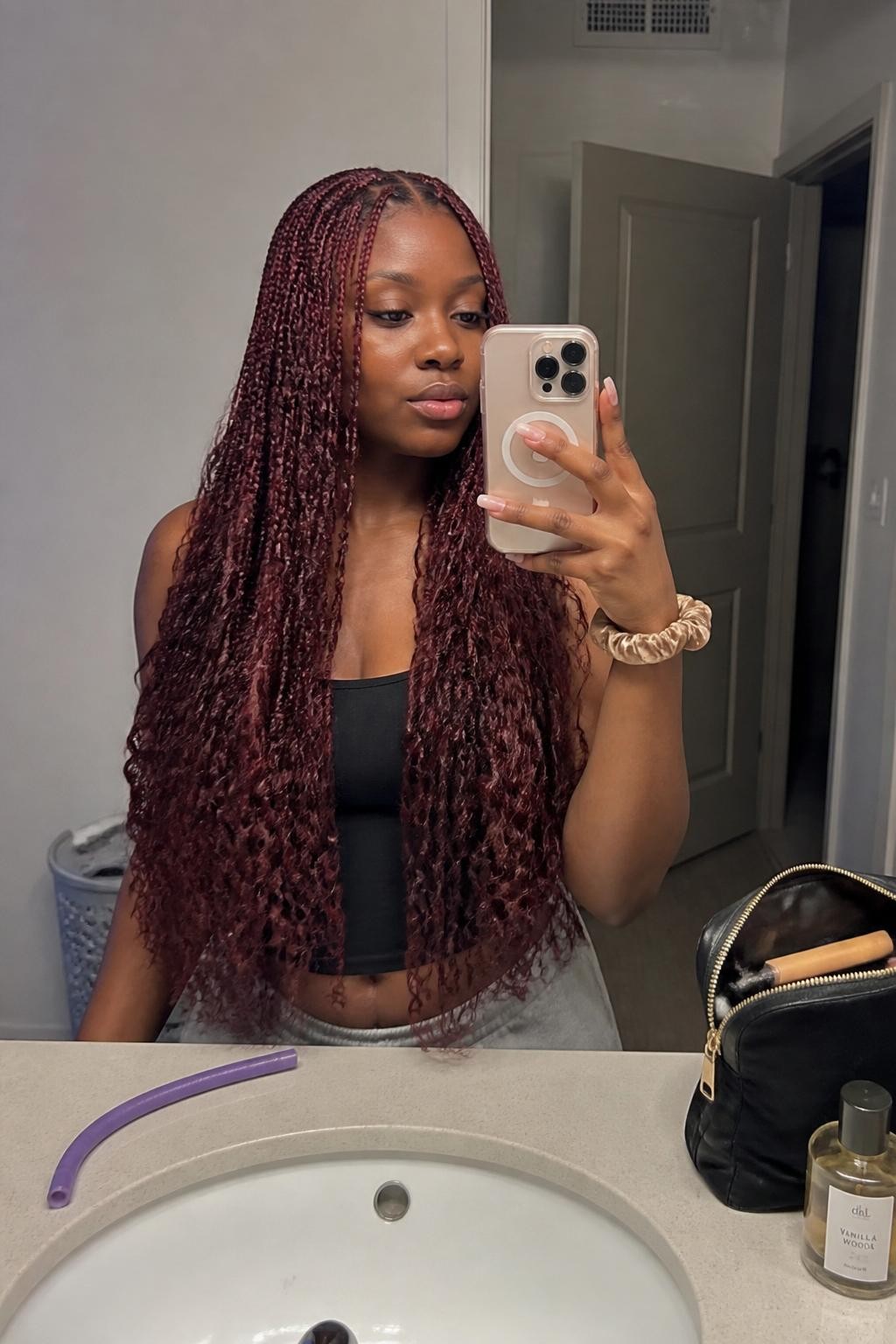 Image prompt: Photo of a Black woman wearing long burgundy boho box braids with loose curls woven through the lengths and a neat center part, candid mirror selfie, bathroom vanity mirror with a makeup bag and perfume bottle nearby, soft indoor lighting, containing a flexi rod on the counter and a silk scrunchie on her wrist, realistic getting-ready moment, latest iPhone photo quality.