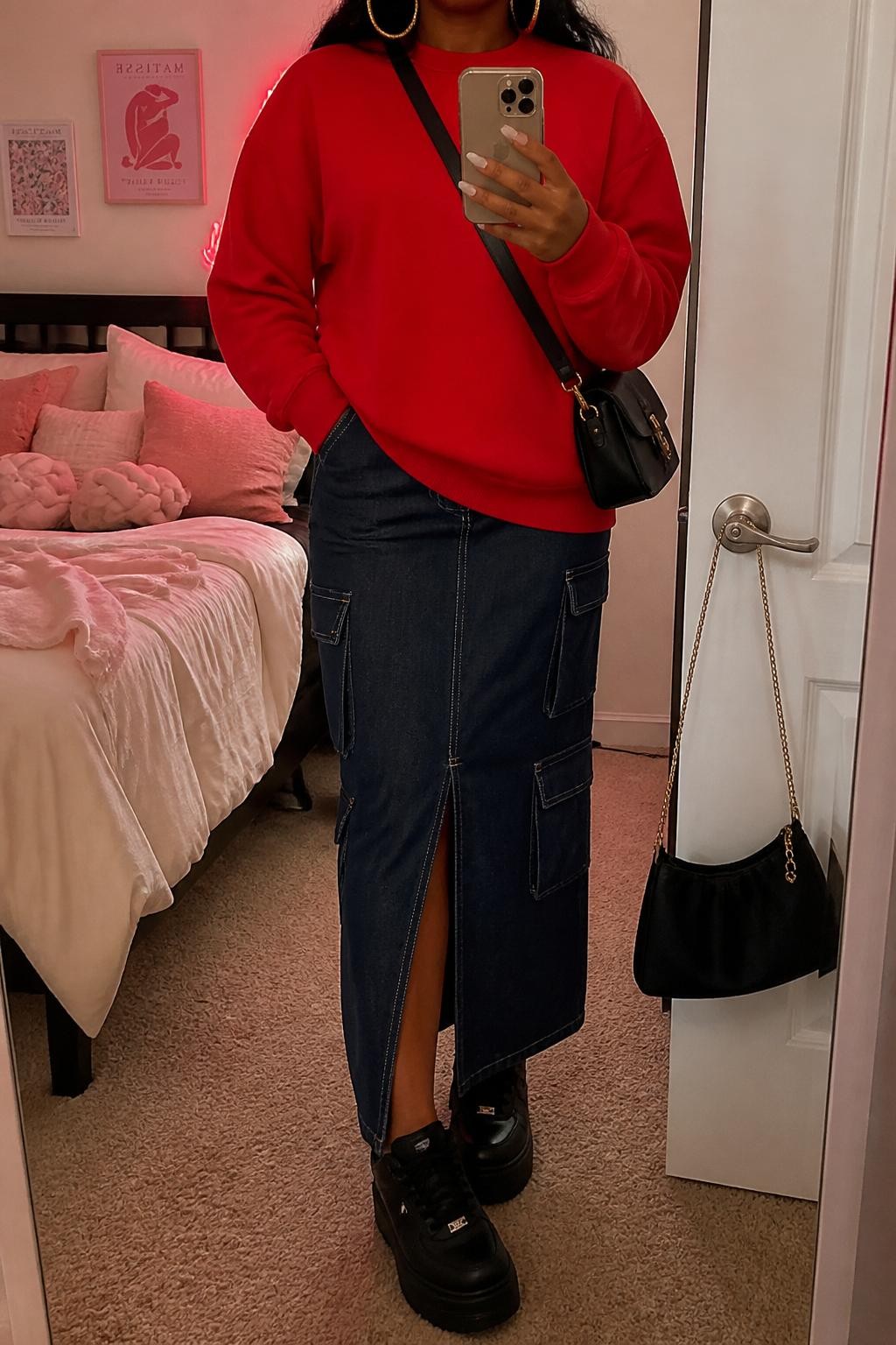 Mirror selfie of a Black woman wearing a red oversized crewneck sweatshirt, dark denim cargo midi skirt with front slit, black platform sneakers, gold hoop earrings, and a black crossbody bag, holding a phone in front of a mirror, cropped at NECK so the face does not show, relaxed casual selfie pose, in a feminine-themed bedroom, warm indoor lighting, with soft pillows on an unmade bed and a handbag hanging on a door handle, latest iPhone photo quality.
