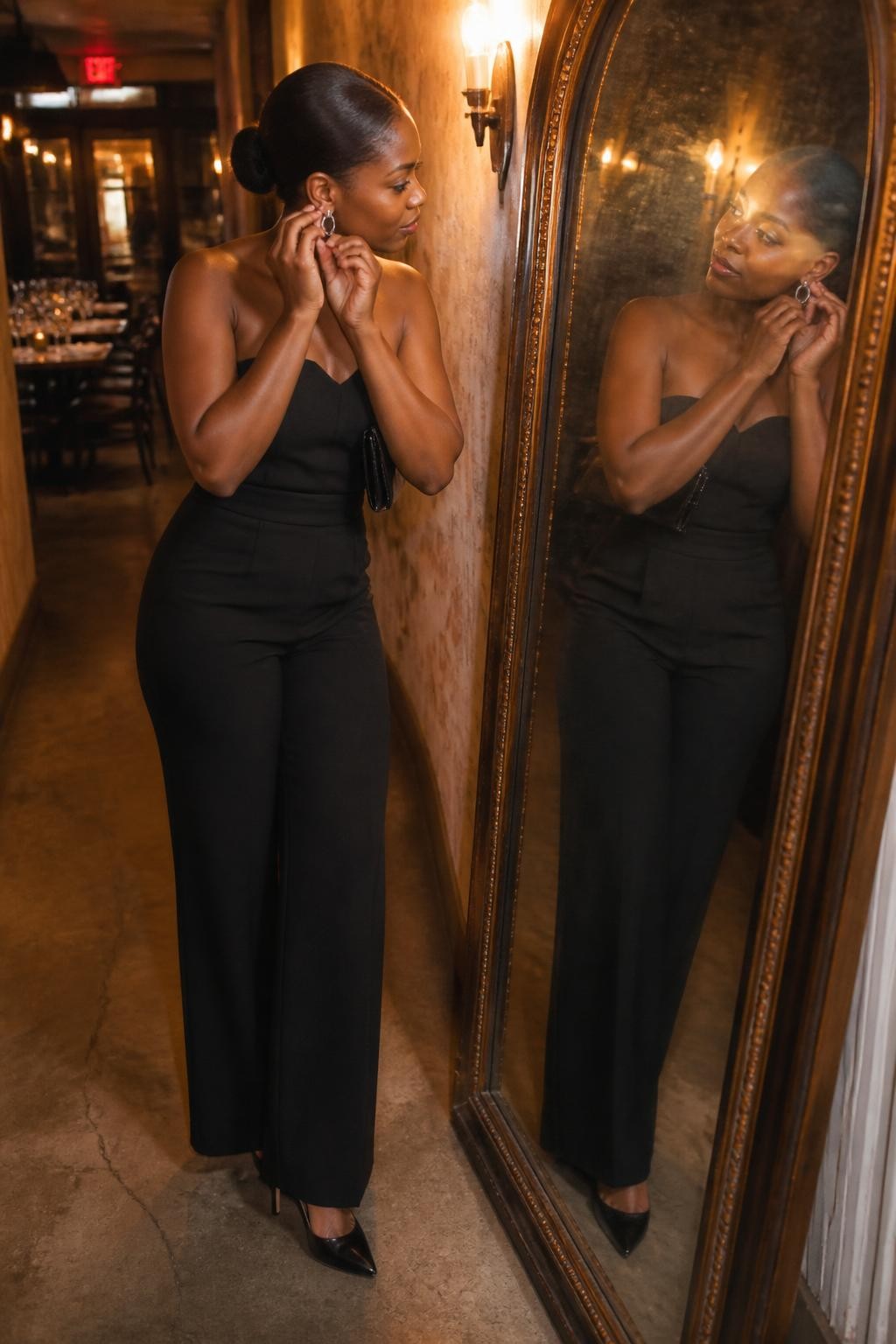 Image prompt: A trendy DSLR restaurant photo of an adult Black woman with warm brown skin standing in a narrow restaurant hallway with a tall antique mirror, wearing a black strapless jumpsuit, pointed heels, a sleek low bun, and silver earrings, adjusting one earring while looking at her reflection, one foot slightly forward, small clutch tucked under her arm, warm wall sconces, textured wallpaper, faint mirror smudges, restaurant tables softly visible behind her through the hallway opening, realistic flash reflection, natural skin texture, stylish night-out mood, human editorial photography.