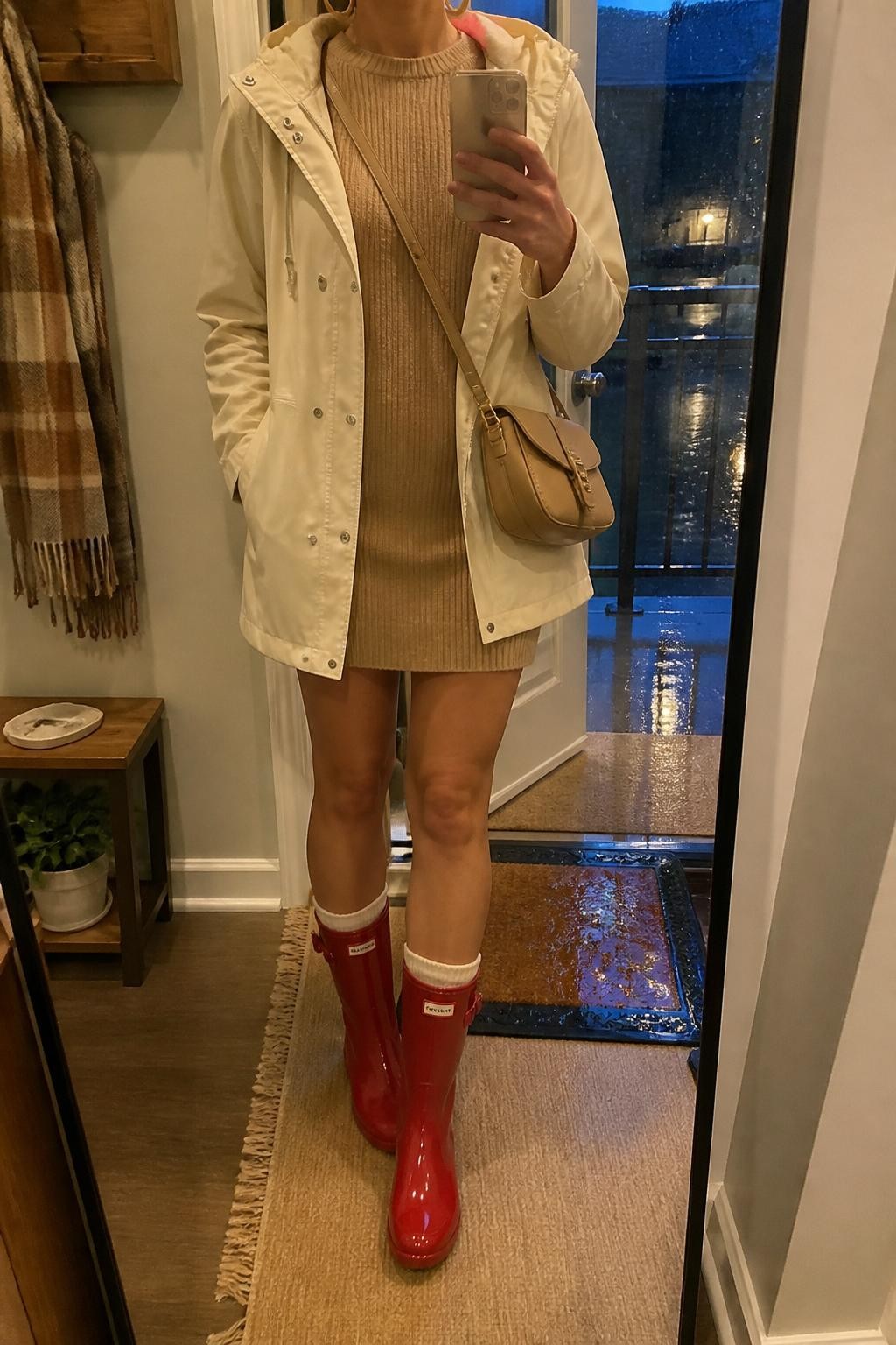 mirror selfie of a woman wearing the exact outfit described above, clearly showing glossy red mid-calf rain boots, a camel ribbed sweater mini dress, cream waterproof jacket worn open, cream crew socks peeking above the boots, a small tan crossbody bag, and gold hoop earrings, holding a phone in front of a mirror, cropped at NECK so the face does not show, with a relaxed casual selfie pose, in a warm apartment entryway mirror, rainy evening light, with a wet doormat near the door and a scarf hanging on a wall hook, latest iPhone photo quality.