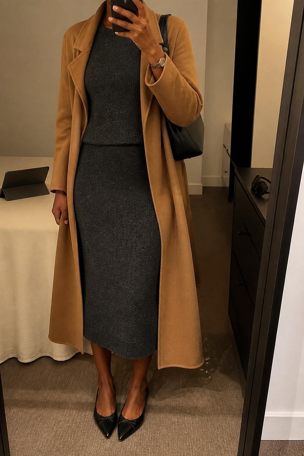 mirror selfie of a Black woman wearing the exact outfit described above, clearly showing a charcoal knit top, matching charcoal knit midi skirt, camel long coat worn open, black pointed flats, a black shoulder bag, and a slim silver watch, holding a phone in front of a mirror, cropped at NECK so the face does not show, with a relaxed casual selfie pose, in a minimal dressing room, warm moody lighting, with a laptop or tablet on the bed and sunglasses placed on a dresser, latest iPhone photo quality.