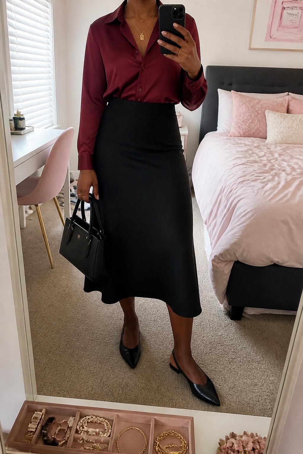 mirror selfie of a Black woman wearing the exact outfit described above, clearly showing a wine red satin blouse tucked into a black A-line midi skirt, black slingback flats, a black structured handbag, and a gold layered necklace, holding a phone in front of a mirror, cropped at NECK so the face does not show, with a relaxed casual selfie pose, in a feminine-themed bedroom, natural window light, with a jewelry tray on the vanity and soft pillows on an unmade bed, latest iPhone photo quality.