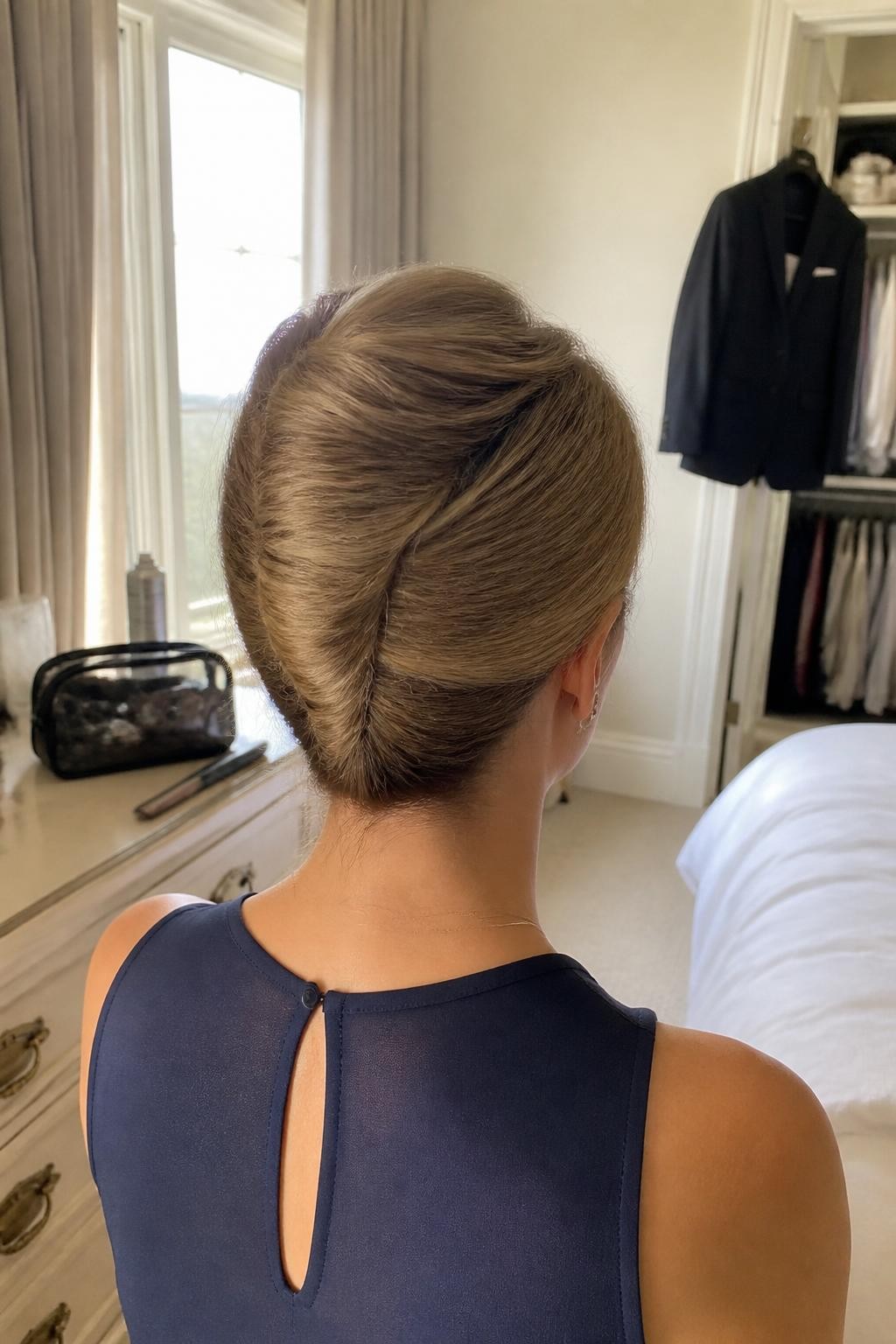 Image prompt: Photo of a woman wearing a polished French twist with soft crown volume and neatly tucked ends, back-of-head view to highlight the upright shape, sun-drenched bedroom with a formal coat hanging near a slightly open closet, natural window light, containing a tail comb on the dresser and a makeup bag nearby, realistic wedding guest styling moment, latest iPhone photo quality.