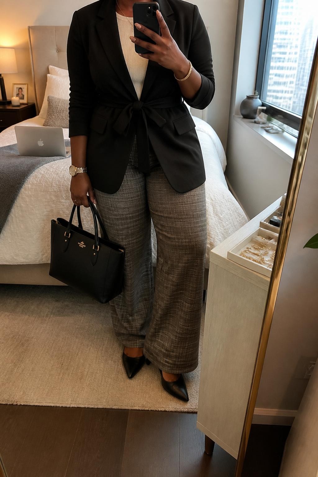 Mirror selfie of a curvy Black woman wearing a black belted blazer, cream fine-knit top, gray and brown plaid wide-leg pants, black pointed flats, and a structured black work tote, holding a phone in front of a mirror, cropped at NECK so the face does not show, relaxed casual selfie pose, in a stylish city apartment, soft indoor lighting, with a laptop on the bed and a jewelry tray on the vanity, latest iPhone photo quality.