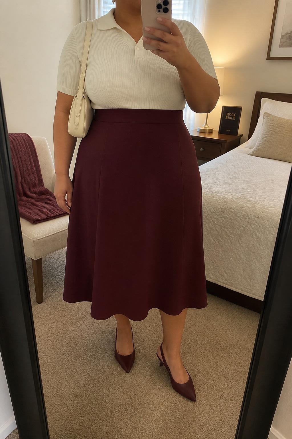 Mirror selfie of a plus size woman wearing a burgundy high-waisted A-line midi skirt in structured fabric, a fitted cream short-sleeve knit polo, burgundy pointed kitten heels, a structured cream shoulder bag, and small gold stud earrings, clearly showing the exact clothing items, burgundy skirt, cream knit, footwear, and accessories listed, holding a phone in front of a mirror, cropped at the neck so her face does not show, relaxed neat selfie stance, in a cozy bedroom mirror setup with soft indoor lighting, a folded blanket on a chair and a Bible resting on a bedside table, realistic church outfit, latest iPhone photo quality.