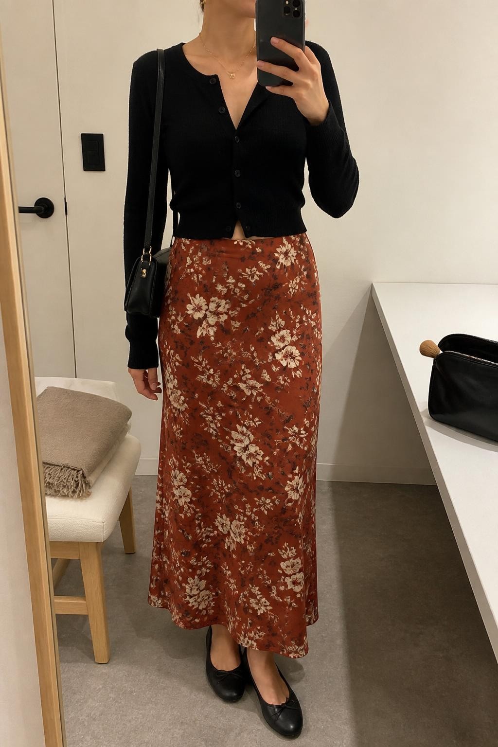 mirror selfie of a woman wearing the exact outfit described above, clearly showing a rust floral maxi skirt with cream and brown flowers, black cropped button-front cardigan, black ballet flats, black mini shoulder bag, gold pendant necklace, and small gold earrings, holding a phone in front of a mirror, cropped at NECK so the face does not show, with a relaxed casual selfie pose, in a minimal dressing room, soft indoor lighting, with folded blanket on a chair and makeup brush or cosmetic bag on the counter, latest iPhone photo quality.