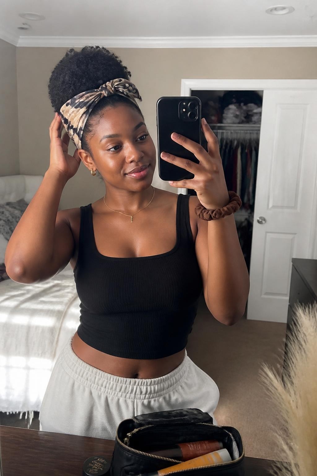 Image prompt: Photo of a Black woman wearing a Curly High Puff with Printed Headband candid mirror selfie, sun-drenched bedroom with a slightly open closet behind her, natural window light, containing a makeup bag on the dresser and a silk scrunchie on her wrist, realistic morning moment, latest iPhone photo quality.