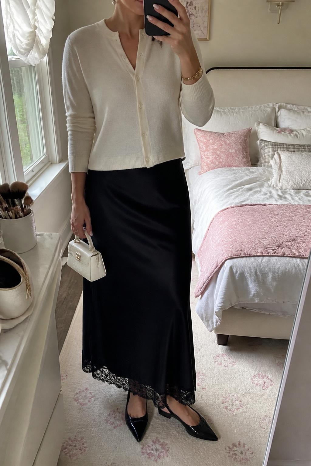 mirror selfie of a woman wearing the exact outfit described above, clearly showing a black lace-trim satin maxi skirt, ivory button-front cardigan, black slingback flats, pearl white mini handbag, pearl stud earrings, and a slim gold watch, holding a phone in front of a mirror, cropped at NECK so the face does not show, with a relaxed casual selfie pose, in a feminine-themed bedroom, natural window light, with makeup brush or cosmetic bag on the counter and soft pillows on an unmade bed, latest iPhone photo quality.