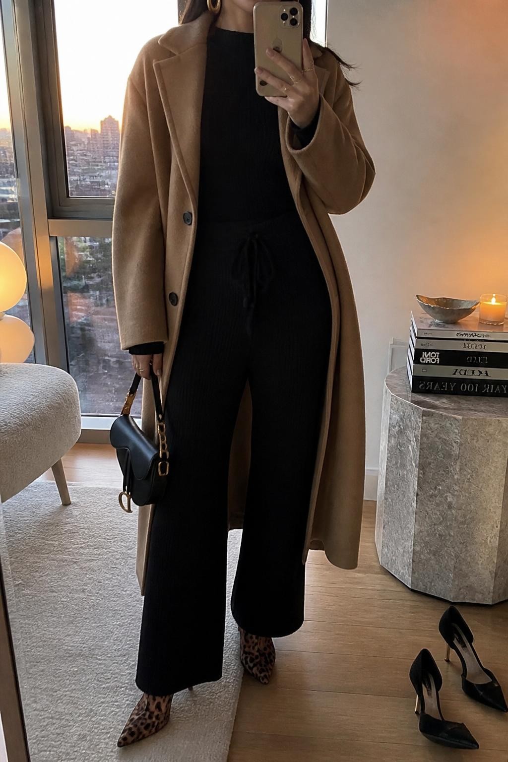 mirror selfie of a woman wearing the exact outfit described above, clearly showing brown leopard animal print ankle boots, a black knit sweater, matching black knit wide-leg pants, a camel long coat, a black mini handbag, and gold hoop earrings, holding a phone in front of a mirror, cropped at NECK so the face does not show, with a relaxed casual selfie pose, in a stylish city apartment, evening golden hour, with a stack of fashion magazines on a side table and a pair of heels on the floor nearby, latest iPhone photo quality.