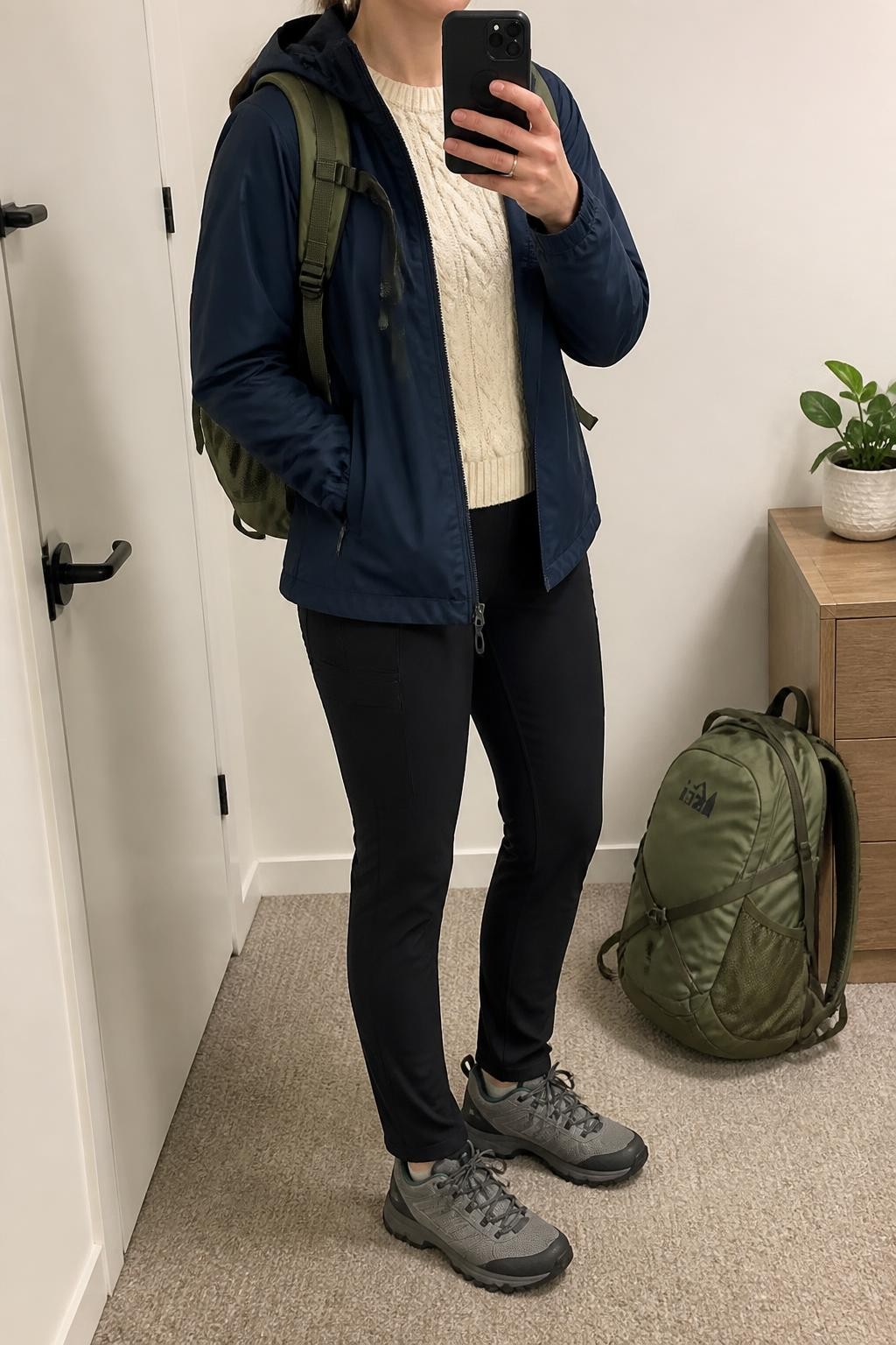 Mirror selfie of a woman wearing a navy water-resistant hooded rain jacket over a cream cable-knit sweater, black straight-leg hiking leggings with side pockets, grey waterproof trail shoes, and an olive-green nylon backpack, clearly showing the exact clothing items, colors, fabrics, and accessories listed, holding a phone in front of a minimal dressing room mirror, cropped at the neck so the face does not show, relaxed casual selfie pose, soft indoor lighting, a backpack resting on the floor and a small plant on a dresser, natural and uncluttered, latest iPhone photo quality.
