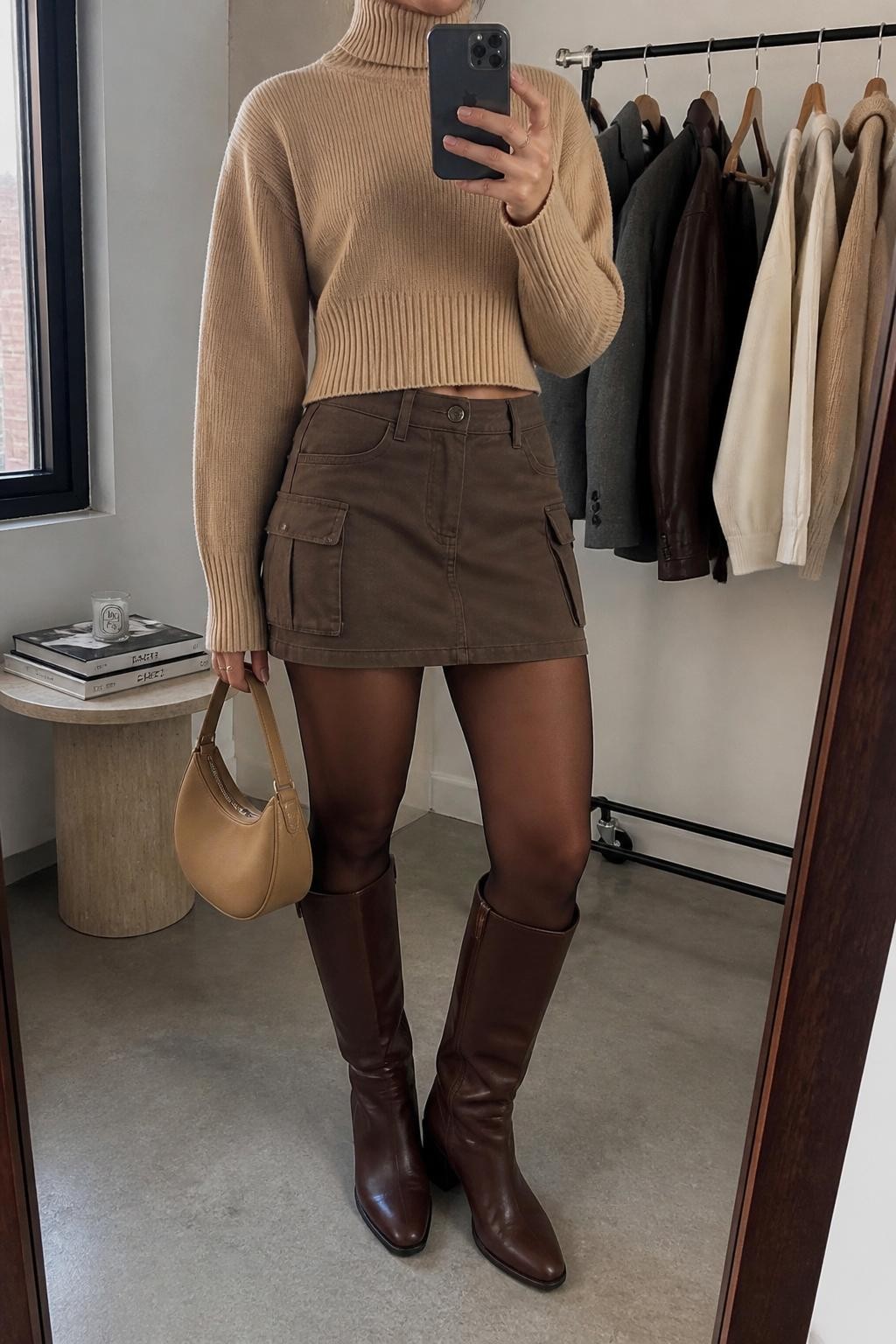 mirror selfie of a woman wearing a camel cropped turtleneck sweater, brown cargo mini skirt, sheer espresso tights, knee-high brown leather boots, and a tan mini hobo bag, clearly showing the same clothing items, colors, fabrics, and accessories listed in the Outfit Pieces section, holding a phone in front of a mirror, cropped at NECK so the face does not show, with a relaxed casual selfie pose, in a minimal dressing room, natural window light, including a clothing rack with garments behind and a stack of fashion magazines on a side table, latest iPhone photo quality.
