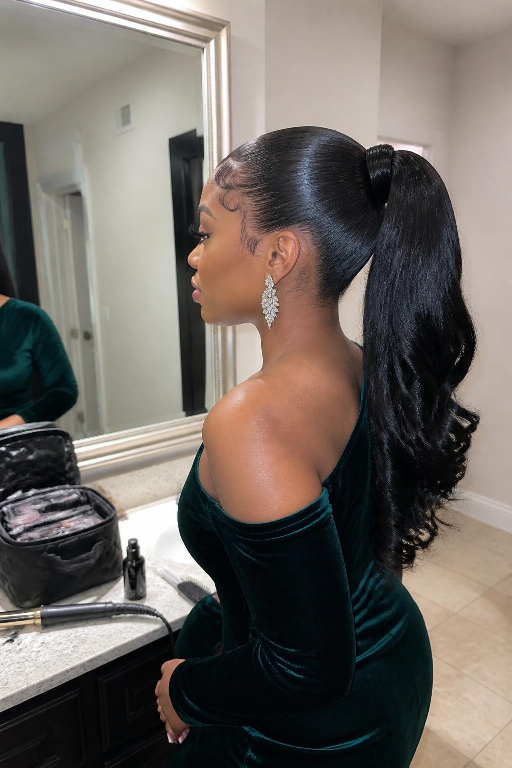 Image prompt: Photo of a Black woman wearing a sleek high ponytail with a wrapped base and softly curled ends, side profile view, bathroom vanity mirror with a formal makeup bag nearby, soft indoor lighting, containing a curling iron on the counter and crystal statement earrings peeking through, realistic fall wedding guest getting-ready moment, latest iPhone photo quality.