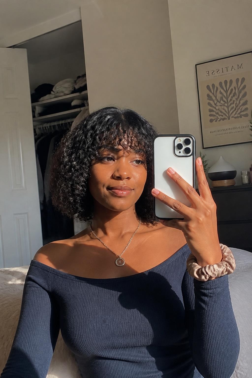 Image prompt: Photo of a Black woman wearing a rounded chin-length curly bob with soft layers and a short curly fringe, candid mirror selfie, sun-drenched bedroom with a slightly open closet and folded sweaters behind her, natural window light, containing a diffuser on the dresser and a silk scrunchie around her wrist, realistic wash-day result, latest iPhone photo quality.