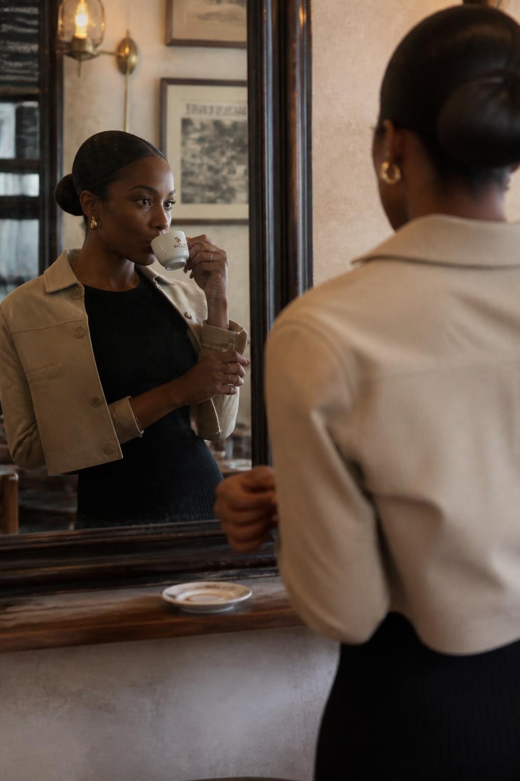 A polished cafe photo of an adult Black woman with rich brown skin reflected in a narrow antique mirror on a cafe wall, wearing a black ribbed midi dress with a cropped beige jacket, standing at a small shelf counter with an espresso cup, one hand lifting the tiny cup near her lips, the other hand adjusting her jacket sleeve, her reflection clear in the mirror while the real scene is partly visible from behind, warm brass lights, framed art, tiny scratches on the mirror glass, dark wood trim, realistic coffee stain on saucer, elegant cafe mood, DSLR editorial style.