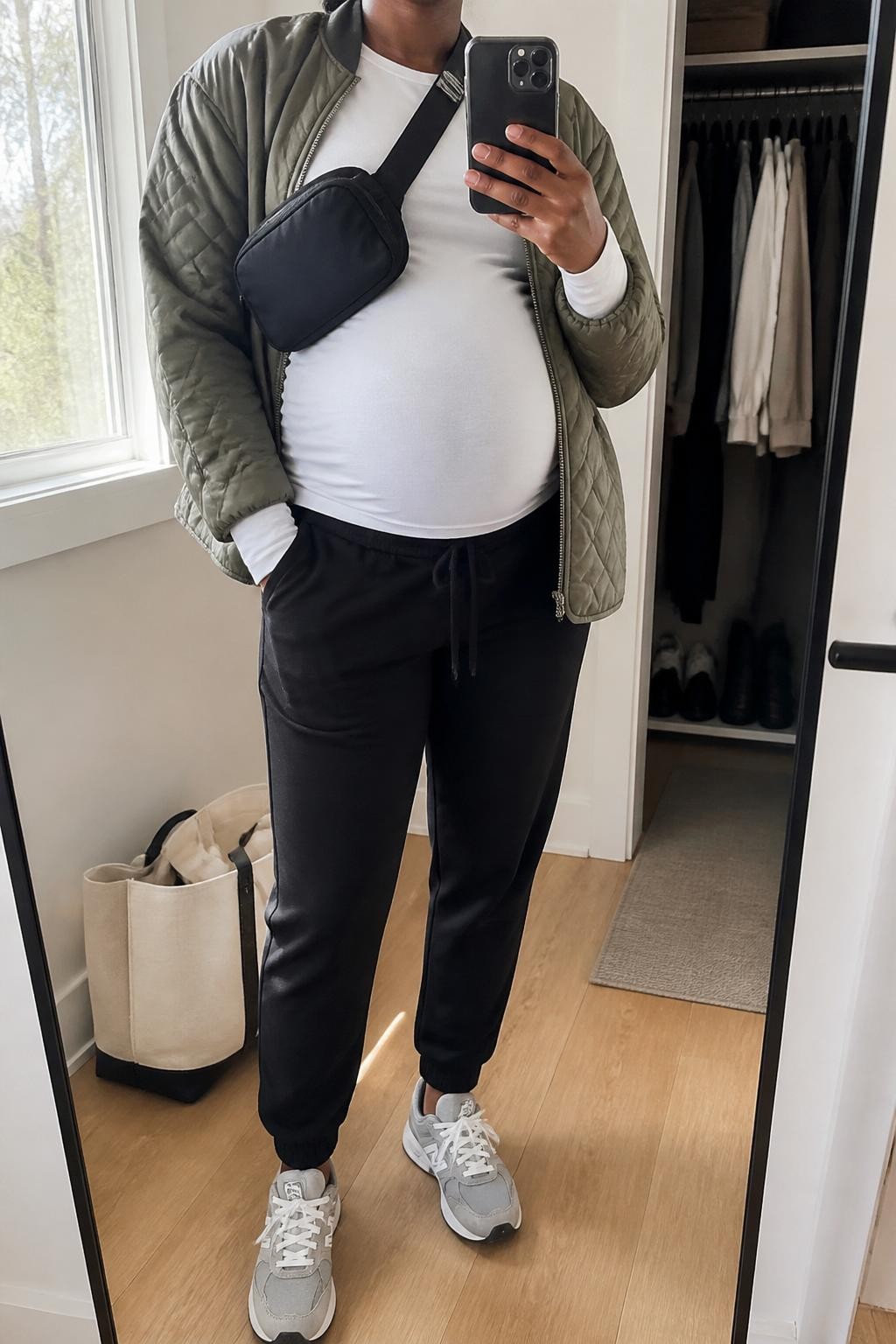 mirror selfie of a pregnant Black woman wearing a fitted white maternity long sleeve tee, black maternity joggers, olive green quilted jacket, gray running sneakers, and a black nylon belt bag, clearly showing the same clothing items, colors, fabrics, and accessories listed in the Outfit Pieces section, holding a phone in front of a mirror, cropped at NECK so the face does not show, with a relaxed casual selfie pose, in a minimal dressing room, natural window light, including a tote bag resting on the floor and an open closet with clothes visible, latest iPhone photo quality.