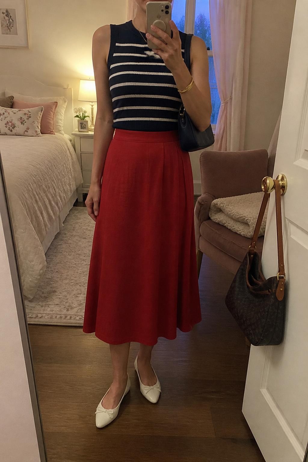 mirror selfie of a woman wearing a fitted navy and white striped sleeveless knit top tucked into a red cotton A-line midi skirt, white pointed ballet flats, navy mini shoulder bag, and gold bangle bracelet, clearly showing the same clothing items, colors, fabrics, and accessories listed in the Outfit Pieces section, holding a phone in front of a mirror, cropped at NECK so the face does not show, with a relaxed casual selfie pose, in a feminine-themed bedroom, evening golden hour, including a folded blanket on a chair and a handbag hanging on a door handle, latest iPhone photo quality.