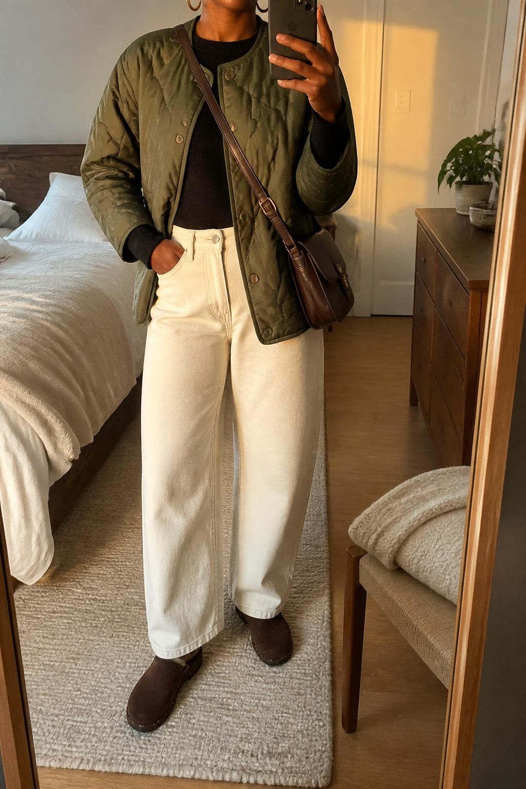 Mirror selfie of a Black woman wearing an olive green quilted collarless jacket over a fitted black long-sleeve top and ecru barrel-leg jeans, clearly showing dark brown suede clogs, a chocolate brown leather crossbody bag, and small gold hoop earrings, holding an iPhone in front of a cozy bedroom mirror setup, cropped at the neck so the face does not show, relaxed casual selfie pose, evening golden hour light, a folded blanket on a chair and a small plant on a dresser, natural unstaged setting, latest iPhone photo quality.
