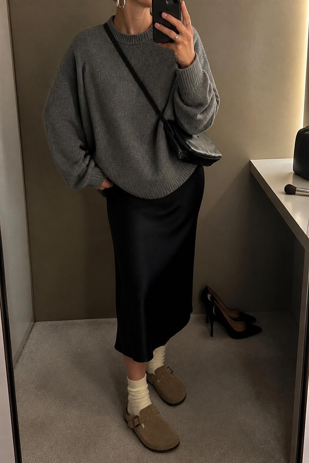 mirror selfie of a woman wearing the exact outfit described above, clearly showing mink suede Birkenstock-style clogs with ivory socks, a black satin midi slip skirt, an oversized gray crewneck sweater, a black leather crossbody bag, and silver hoop earrings, holding a phone in front of a mirror, cropped at NECK so the face does not show, with a relaxed casual selfie pose, in a minimal dressing room, warm moody lighting, with a pair of heels on the floor nearby and a makeup brush or cosmetic bag on the counter, latest iPhone photo quality.