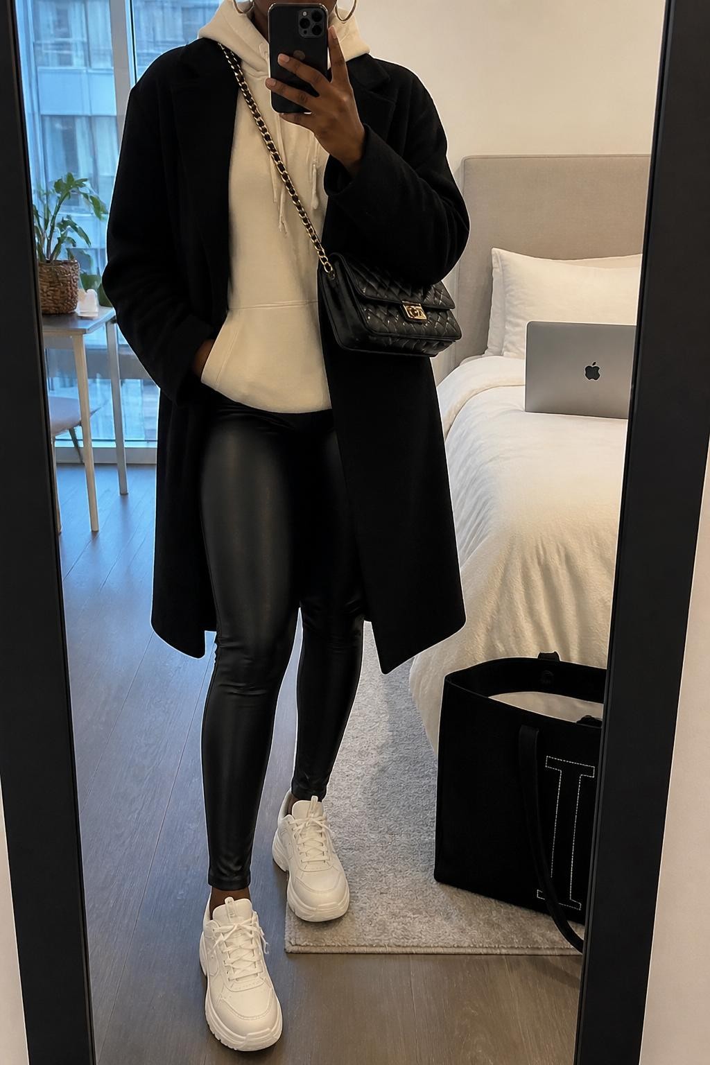 Mirror selfie of a Black woman wearing a black longline coat, cream hoodie, black faux leather leggings, white platform sneakers, gold hoop earrings, and a black quilted crossbody bag, holding a phone in front of a mirror, cropped at NECK so the face does not show, relaxed casual selfie pose, in a stylish city apartment, soft indoor lighting, with a laptop on the bed and a tote bag resting on the floor, latest iPhone photo quality.