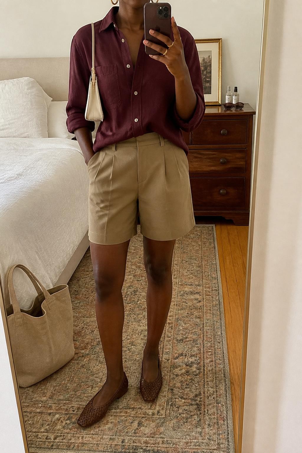 mirror selfie of a Black woman wearing the exact outfit described above, clearly showing a lightweight burgundy cotton button-down shirt half-tucked into tailored tan Bermuda shorts, brown woven flats, a cream mini handbag, and gold hoop earrings, holding a phone in front of a mirror, cropped at NECK so the face does not show, with a relaxed casual selfie pose, in a cozy bedroom mirror setup, warm natural light, with sunglasses on a dresser and a tote bag resting near the bed, latest iPhone photo quality.