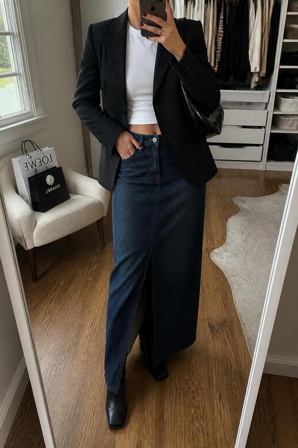 mirror selfie of a woman wearing a black pinstripe fitted blazer, white baby tee, dark blue denim maxi skirt with a front slit, black square-toe boots, and a black patent shoulder bag, clearly showing the same clothing items, colors, fabrics, and accessories listed in the Outfit Pieces section, holding a phone in front of a mirror, cropped at NECK so the face does not show, with a relaxed casual selfie pose, in a walk-in closet mirror, natural window light, including an open closet with clothes visible and shopping bags beside a chair, latest iPhone photo quality.