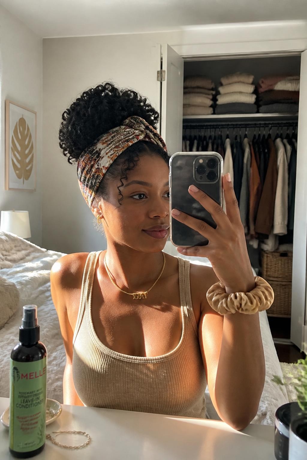 Image prompt: Photo of a Black woman wearing a high curly pineapple updo wrapped with an olive, rust, and cream printed scarf, candid mirror selfie, sun-drenched bedroom with a slightly open closet and folded sweaters behind her, natural window light, containing a curl spray bottle on the dresser and a silk scrunchie on her wrist, realistic relaxed morning moment, latest iPhone photo quality.