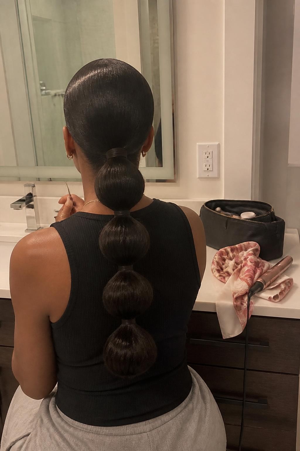 Image prompt: Photo of a Black woman wearing a sleek low bubble ponytail with four soft rounded sections and a clean middle part, back-of-head view to highlight the details, bathroom vanity mirror with a makeup bag nearby, soft indoor lighting, containing a curling iron on the counter and a silk scarf draped beside the sink, realistic getting-ready moment, latest iPhone photo quality.