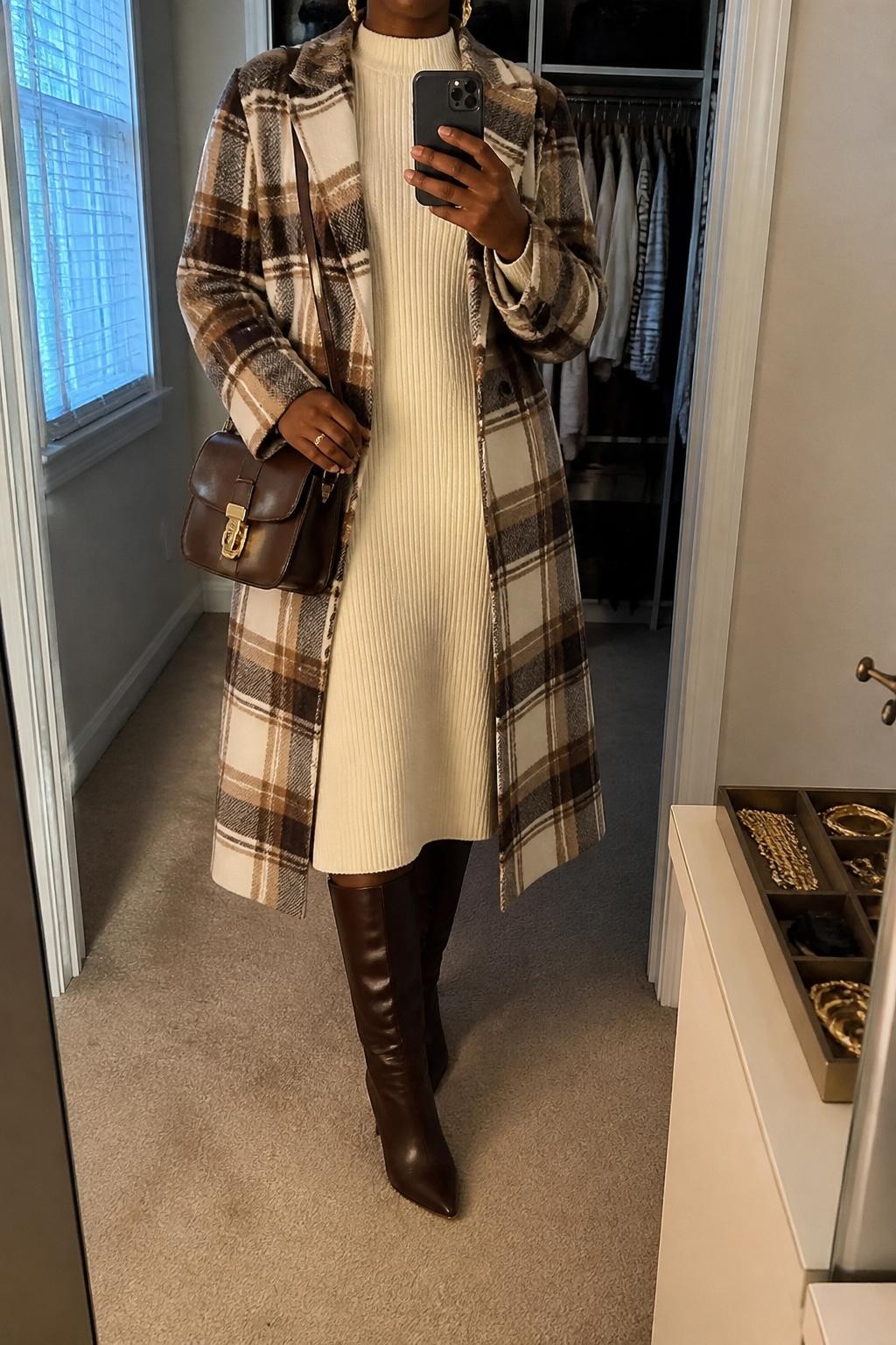 mirror selfie of a Black woman wearing the exact outfit described above, clearly showing a brown and cream plaid longline coat layered over an ivory sweater midi dress, dark brown knee-high boots, a brown leather satchel bag, and gold drop earrings, holding a phone in front of a mirror, cropped at NECK so the face does not show, with a relaxed casual selfie pose, in a walk-in closet mirror, natural window light, with an open closet with clothes visible and a jewelry tray on the vanity, latest iPhone photo quality.